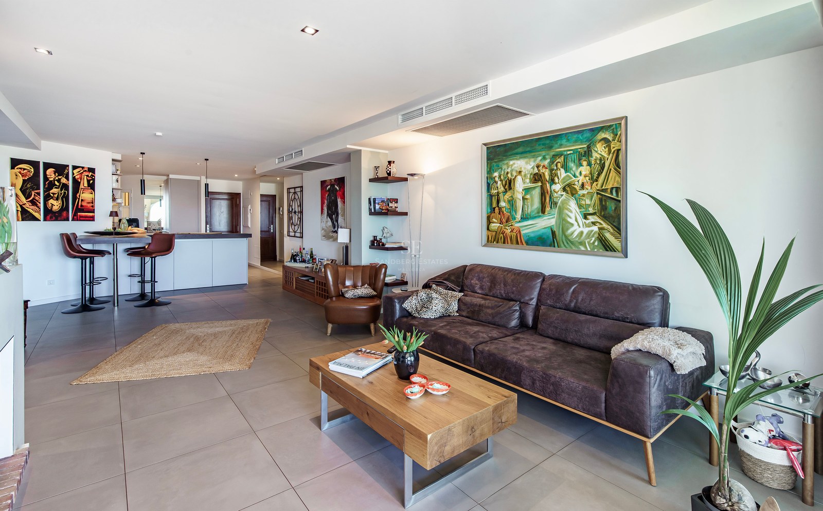 Modern living room featuring a brown leather sofa, wooden coffee table, large-format grey tiles, and integrated kitchen.