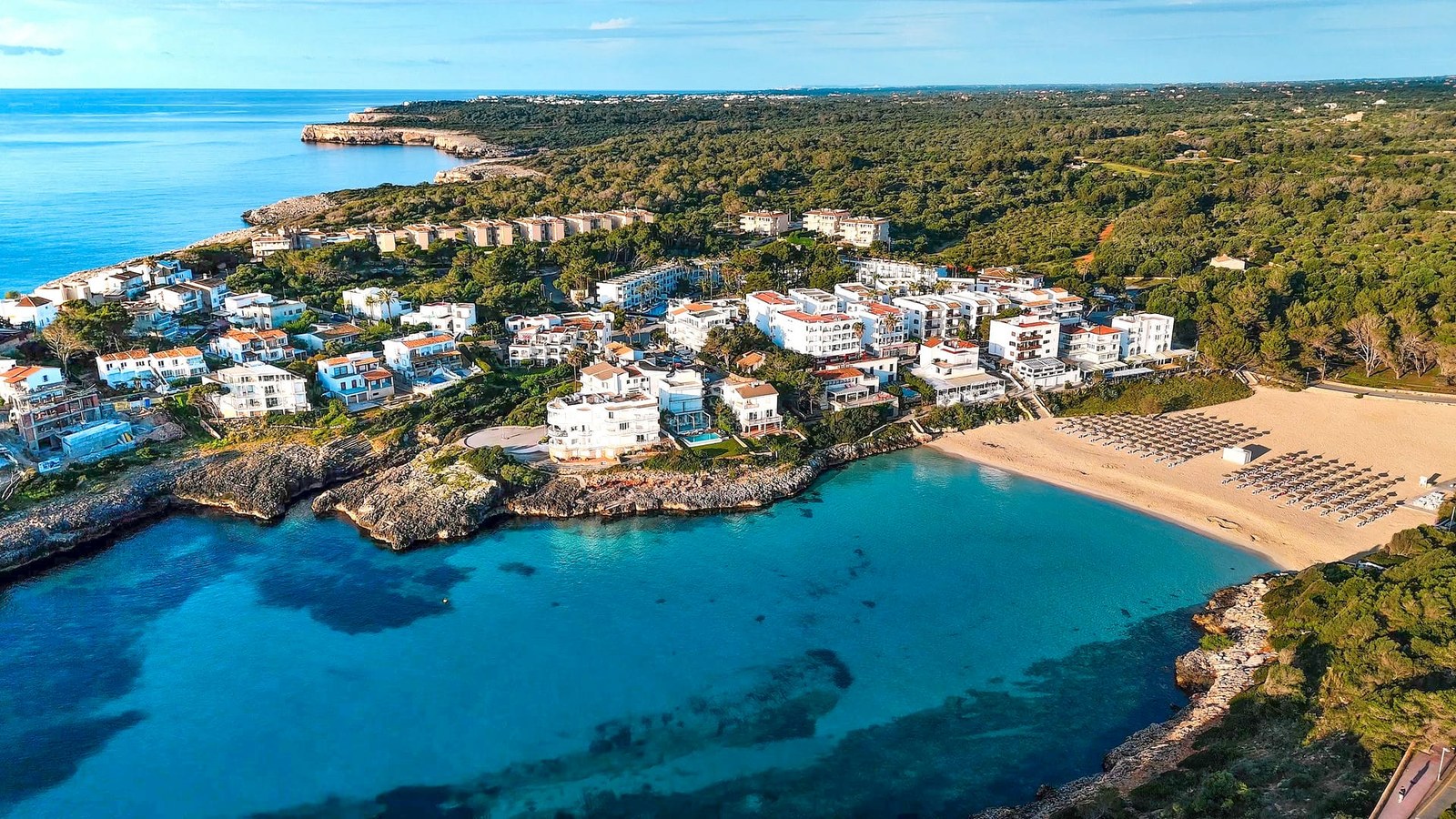 Aerial drone view of a turquoise bay with a sandy beach surrounded by white Mediterranean buildings and lush green forest.