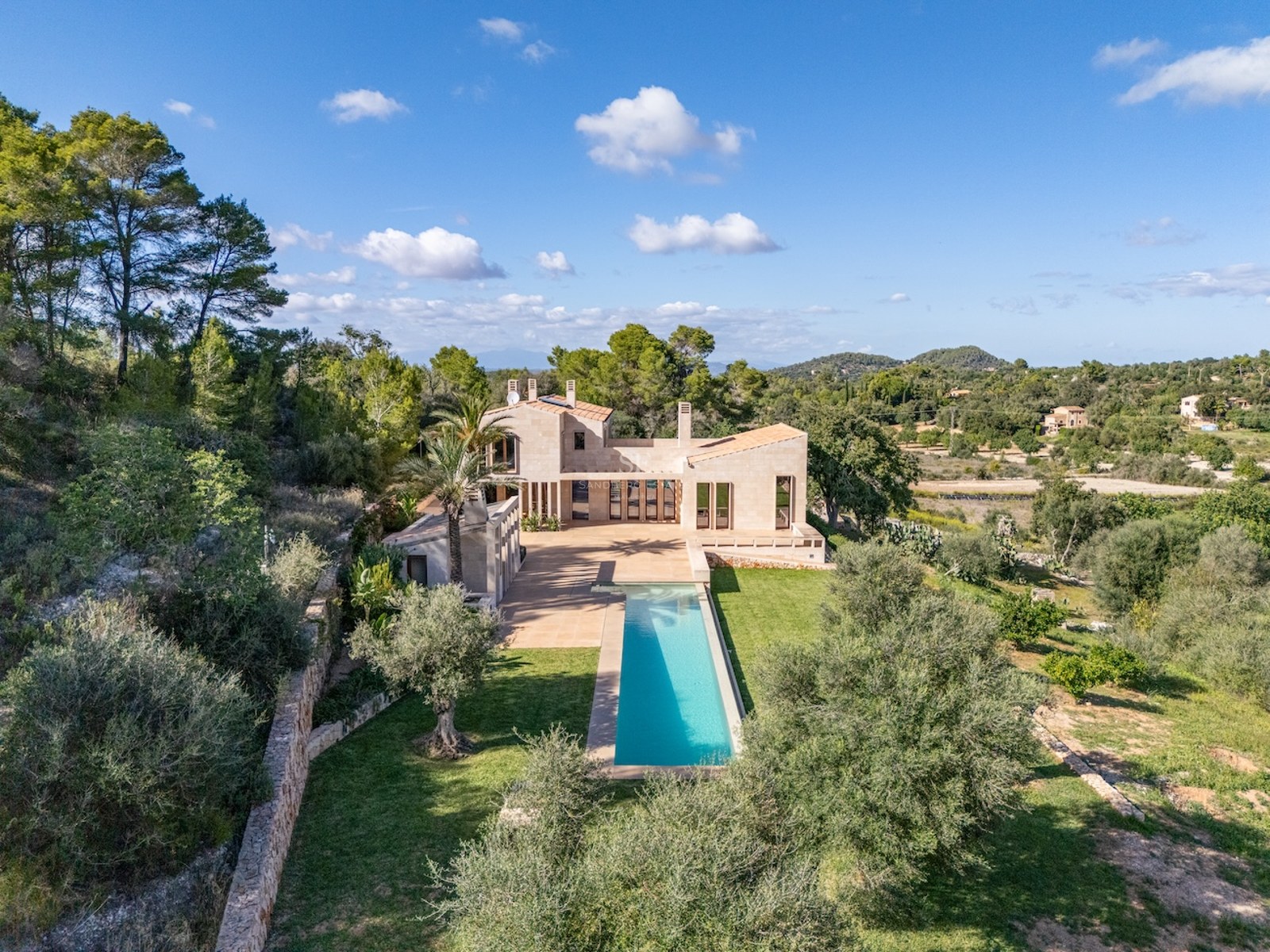 Aerial-style view of a modern stone villa with a long turquoise lap pool surrounded by lush Mediterranean trees.