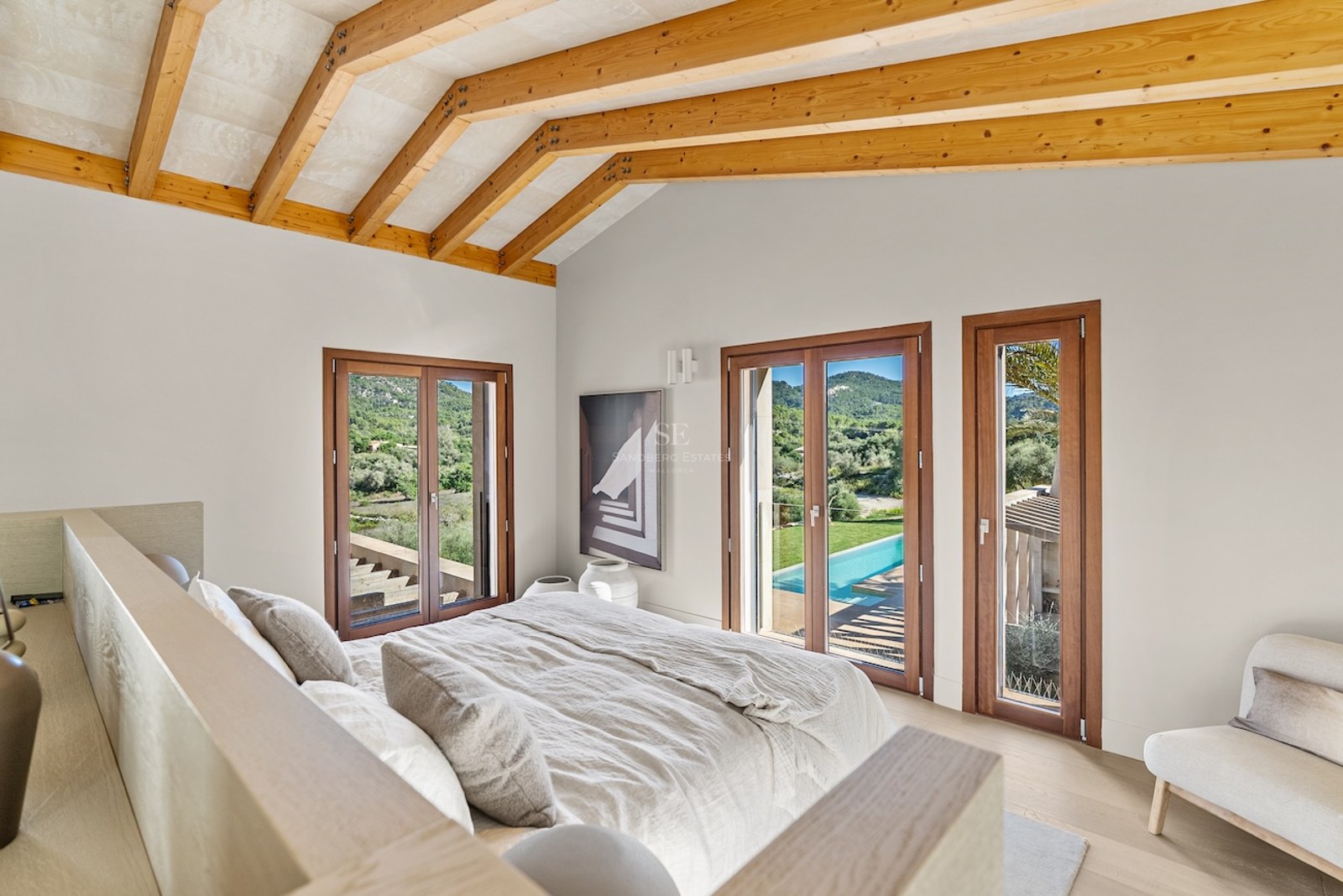 Modern bedroom with vaulted wood beam ceiling, bed with beige linens, and glass doors overlooking a pool and green hills.