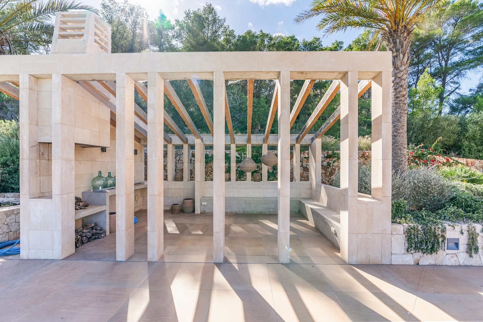 Modern stone pavilion with wooden rafters, outdoor fireplace, and built-in seating surrounded by lush greenery.