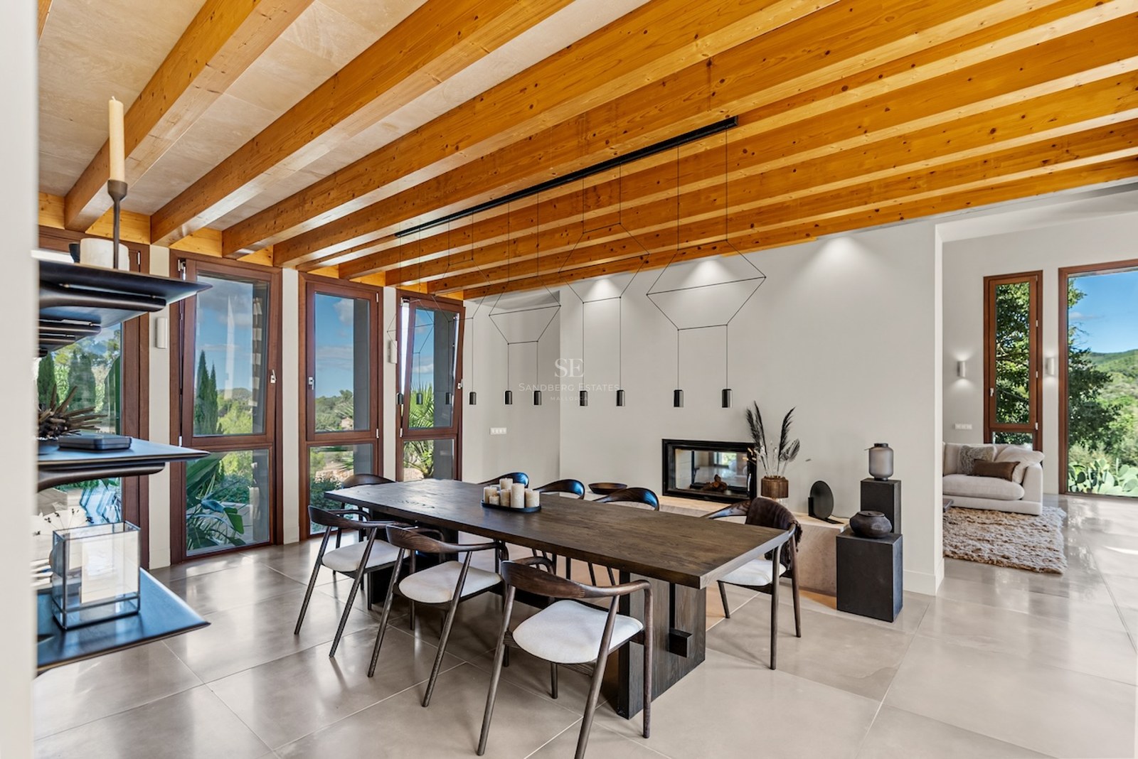 Large wooden dining table under a beamed ceiling with floor-to-ceiling windows showing nature views.