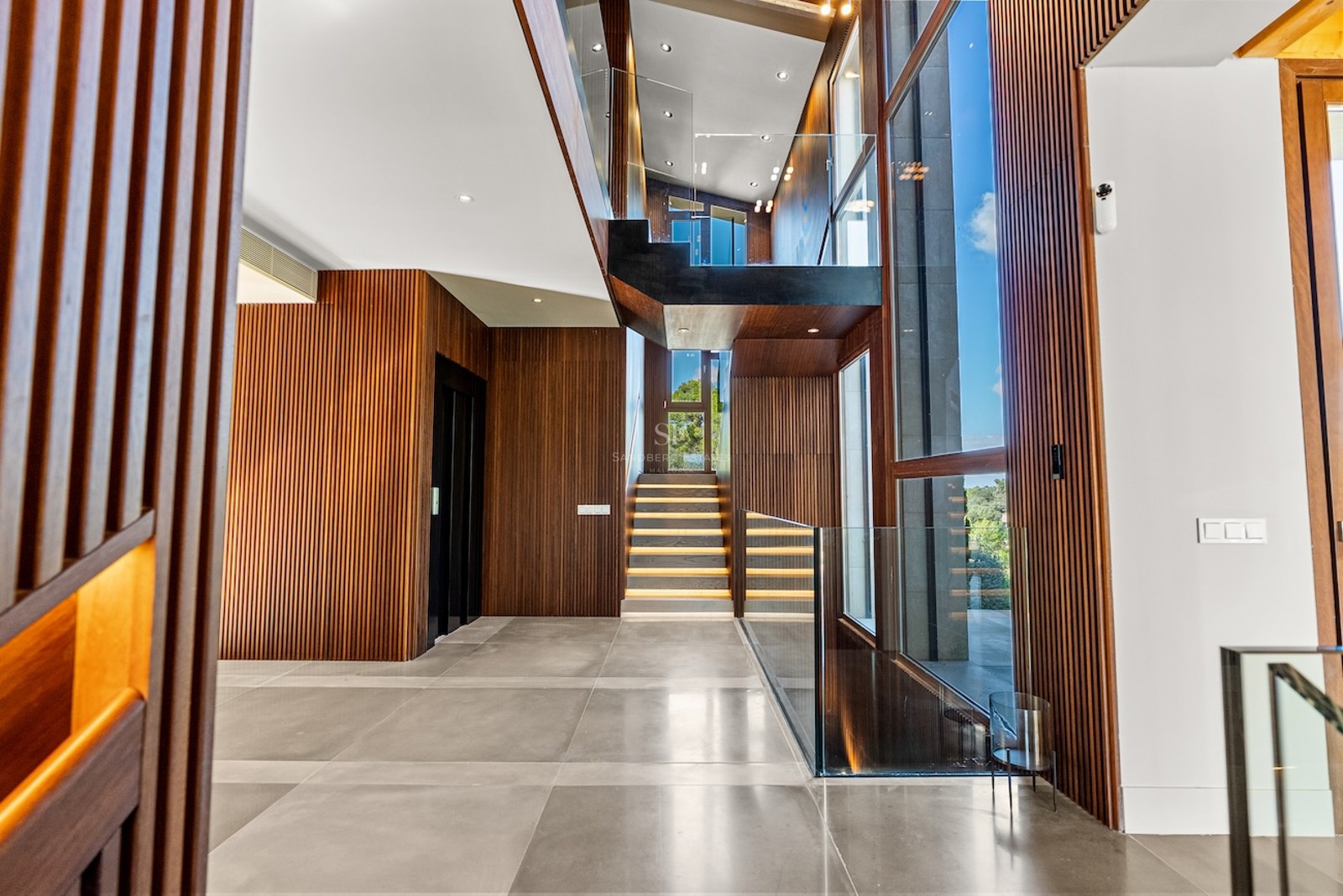 Modern foyer featuring slatted wood walls, a glass-railed staircase with LED lighting, and massive floor-to-ceiling windows.