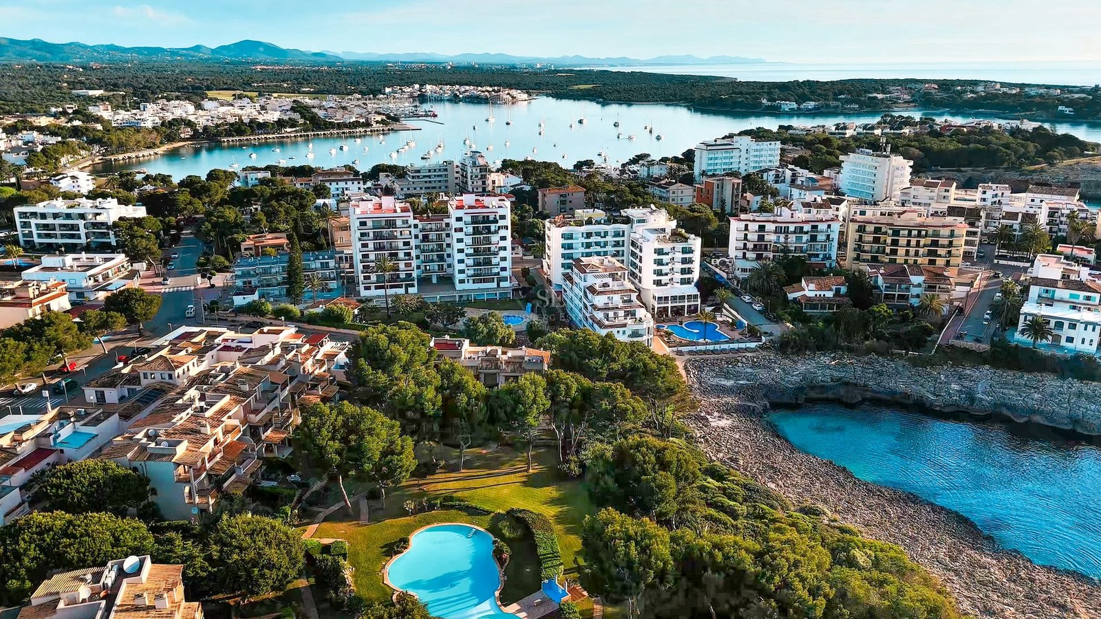 Drone view of a Mediterranean coastal town with white apartment buildings, swimming pools, and a blue bay with boats.