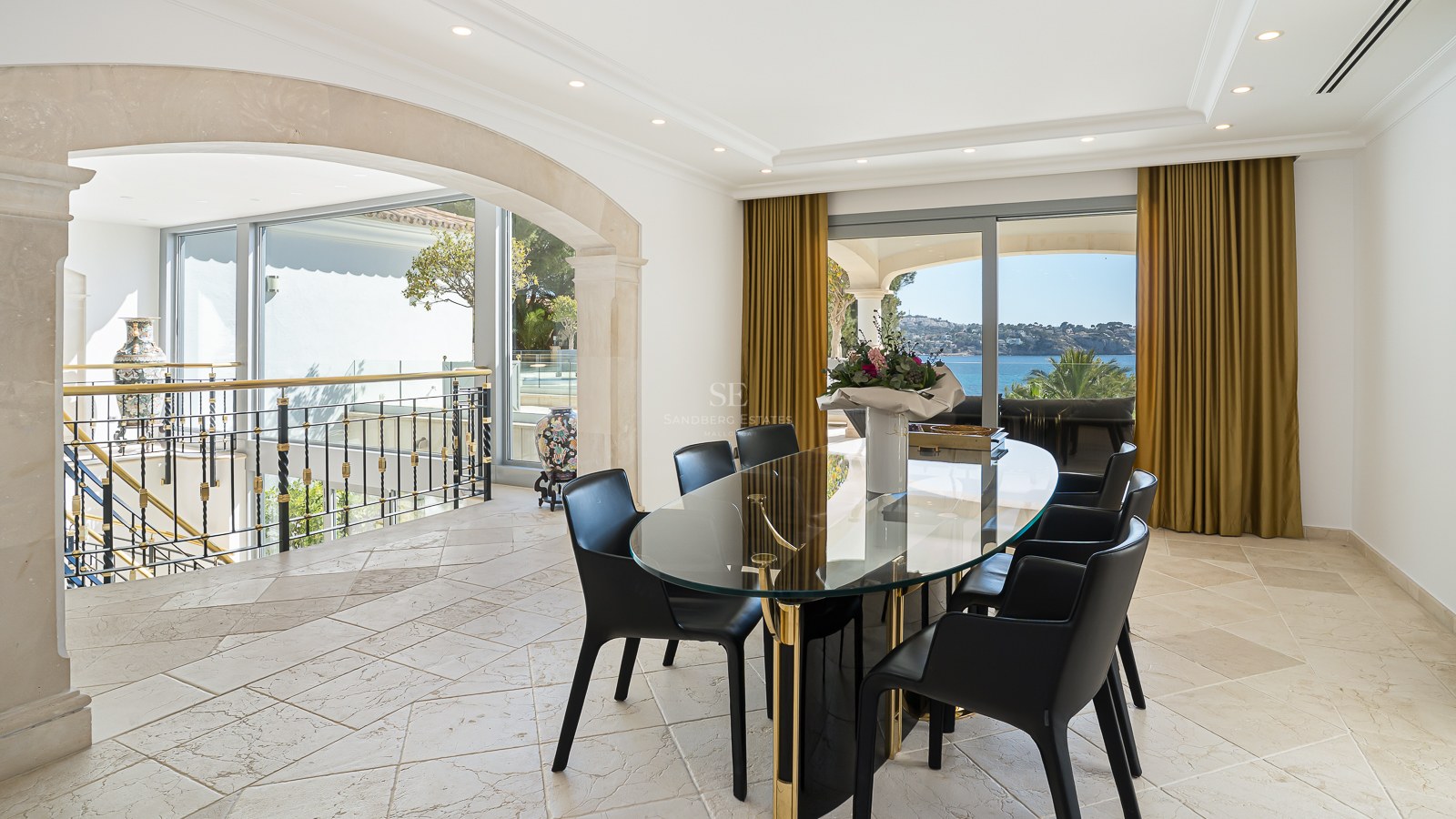 Salle à manger moderne avec table ovale en verre, chaises noires et grandes baies vitrées donnant sur la mer.