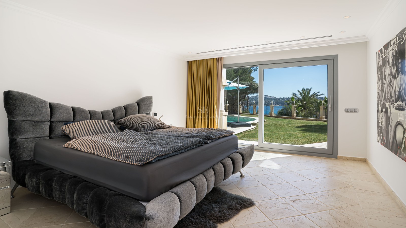 Chambre moderne avec lit gris capitonné et grandes baies vitrées ouvrant sur un jardin, une piscine et la mer.