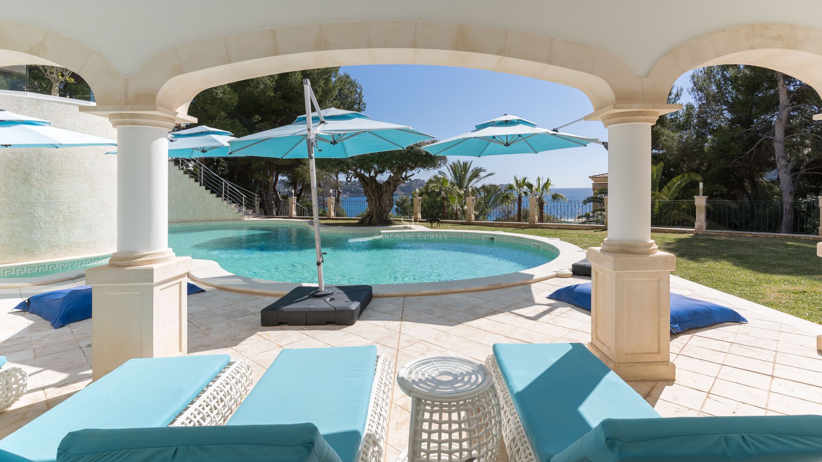 Une piscine turquoise incurvée entourée de dalles en pierre et de chaises longues turquoises sous une arche ombragée.