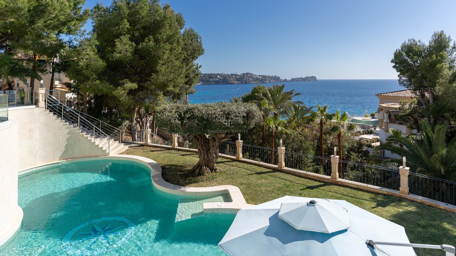 Piscine incurvée turquoise entourée de pelouse et d'oliviers surplombant la mer Méditerranée bleue.