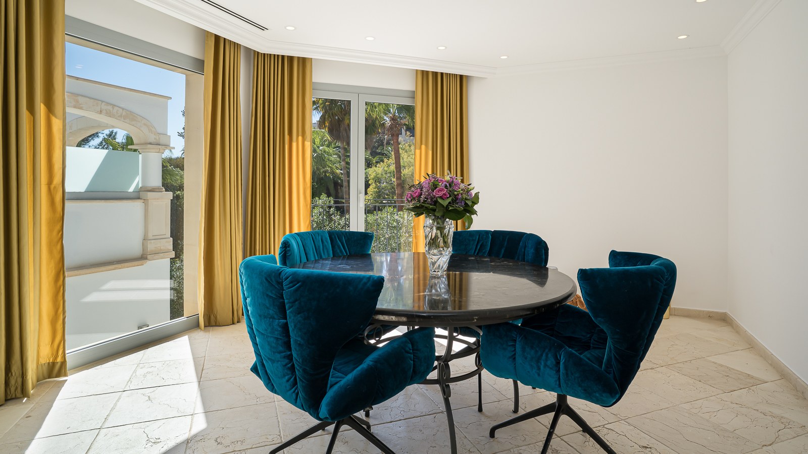 Salle à manger moderne avec table ronde en marbre, chaises en velours bleu et vue sur le jardin.