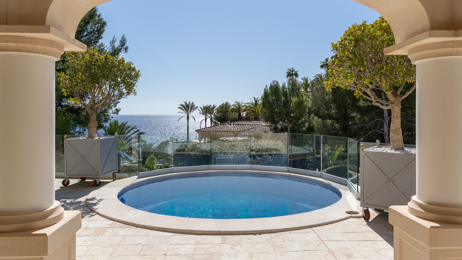 Piscine circulaire bleue sur une terrasse en pierre avec garde-corps en verre offrant une vue sur la mer.