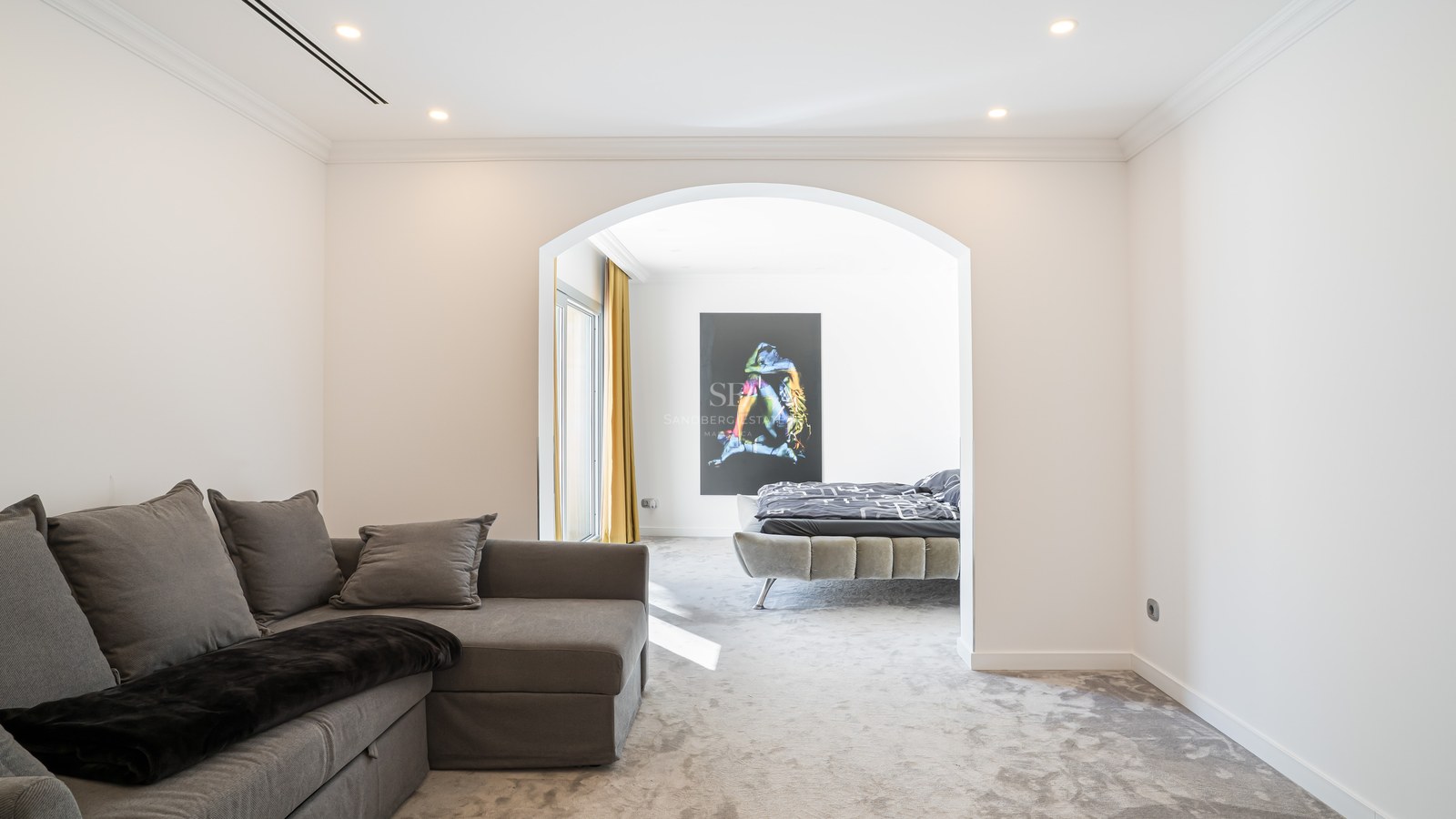 Chambre moderne vue à travers une arche avec lit gris tapissé, moquette épaisse et grande œuvre d'art murale.