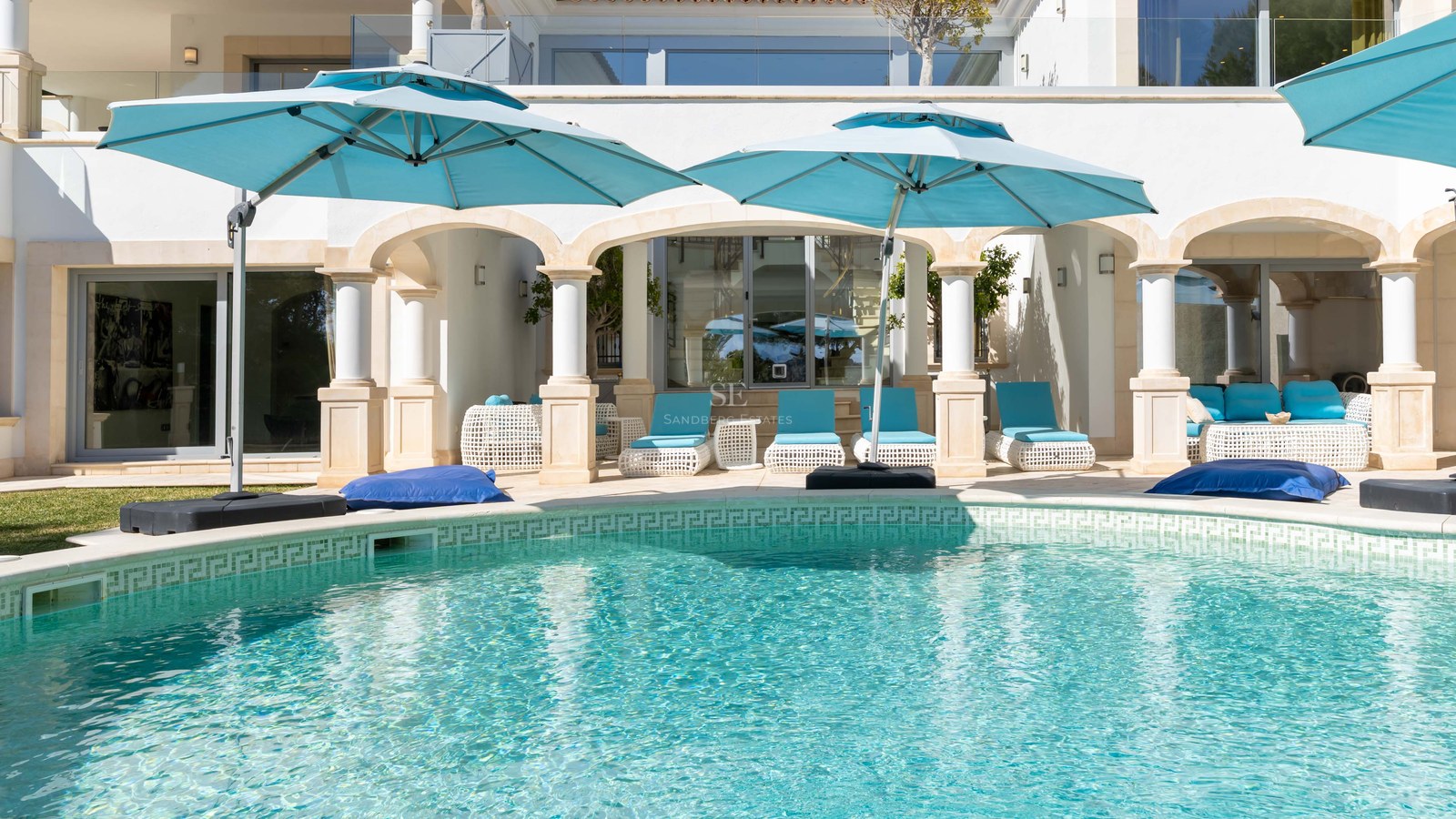 Piscine turquoise avec colonnes blanches classiques et parasols bleu canard sur une terrasse ensoleillée.