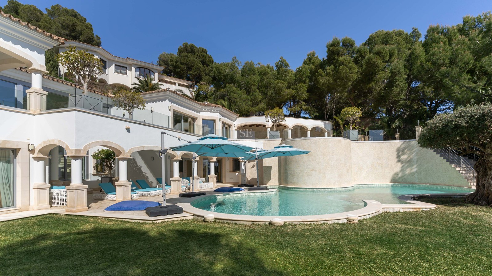 Piscine incurvée avec parasols turquoise à côté d'une villa blanche méditerranéenne entourée de pins.