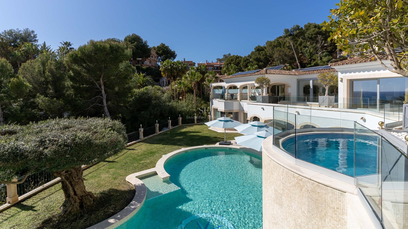 Villa de luxe avec piscine turquoise incurvée, garde-corps en verre et végétation dense à Majorque.