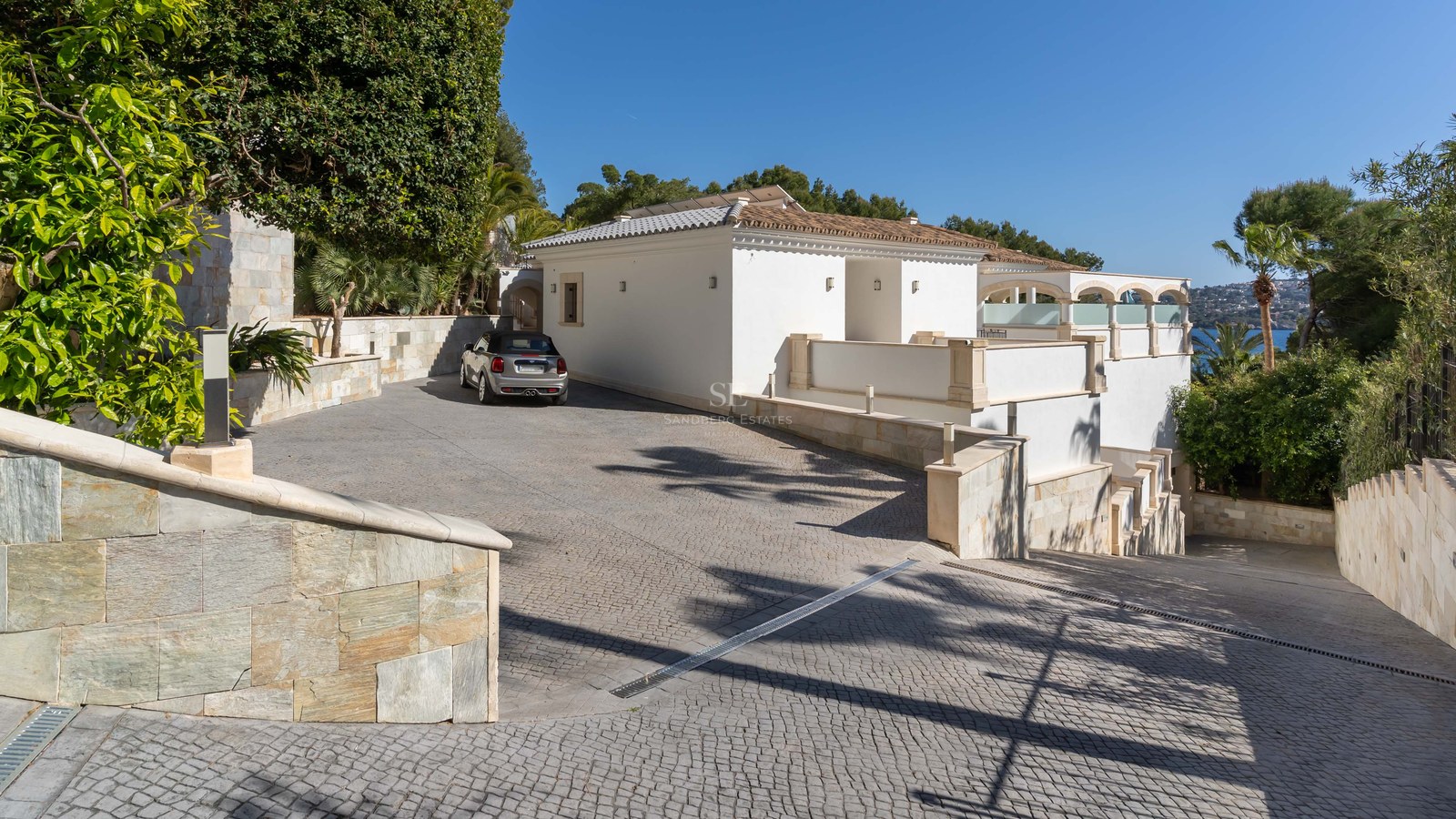 Villa méditerranéenne blanche avec allée pavée privée, murs en pierre et voiture garée sous un ciel bleu.