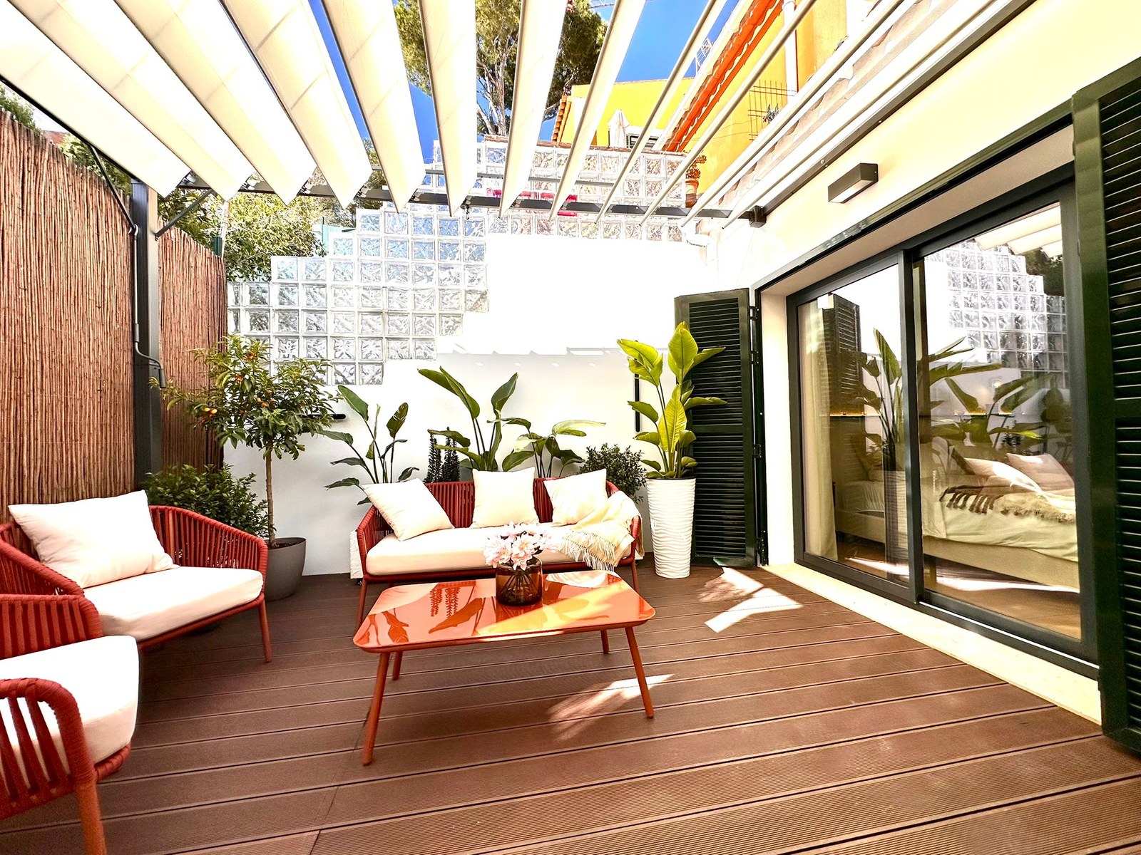 Stylish outdoor terrace with red and white furniture, orange table, lush plants, and a white retractable awning in the sun.