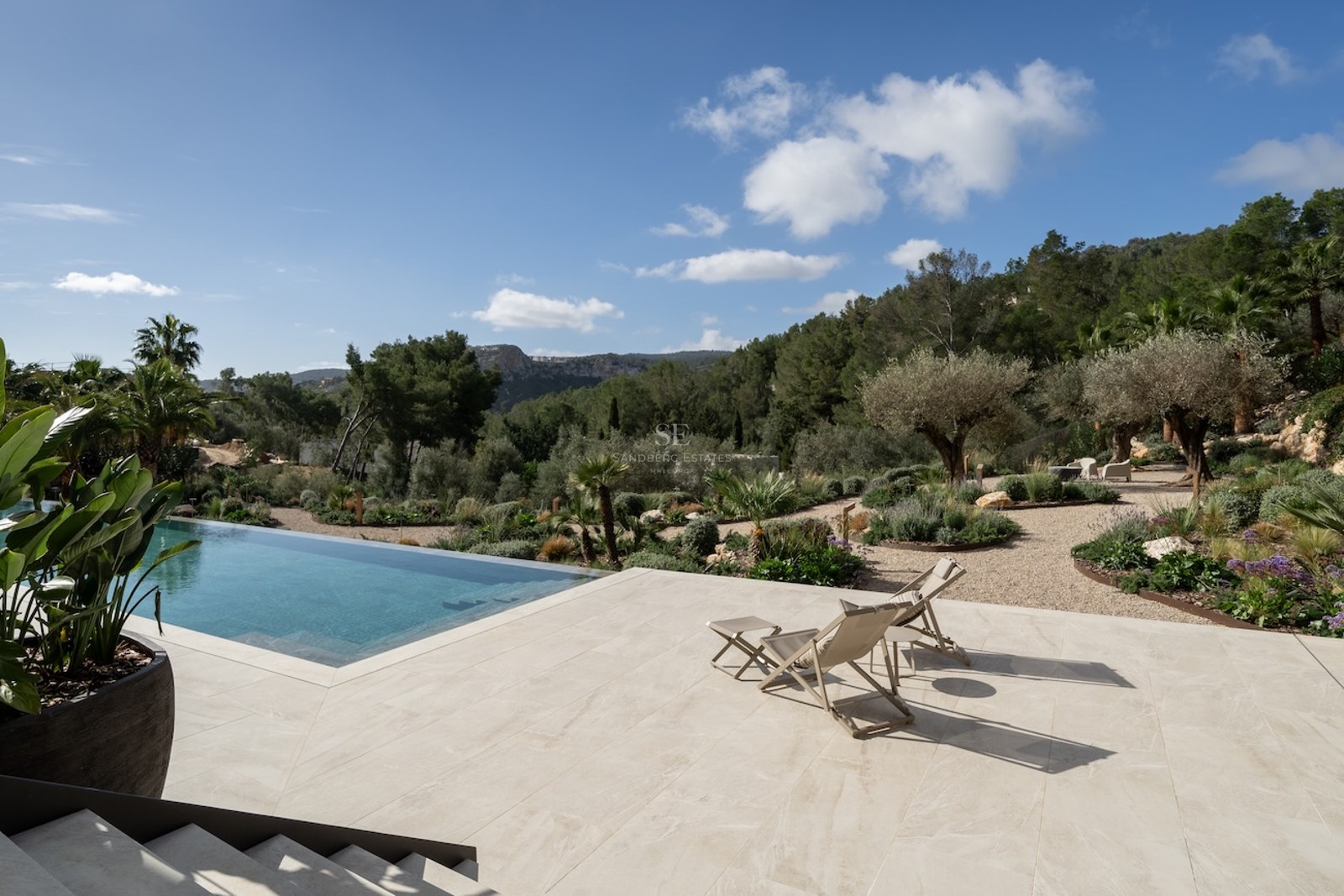 Piscina infinita y terraza de piedra con tumbonas frente a un paisaje mediterráneo de montañas y olivos.