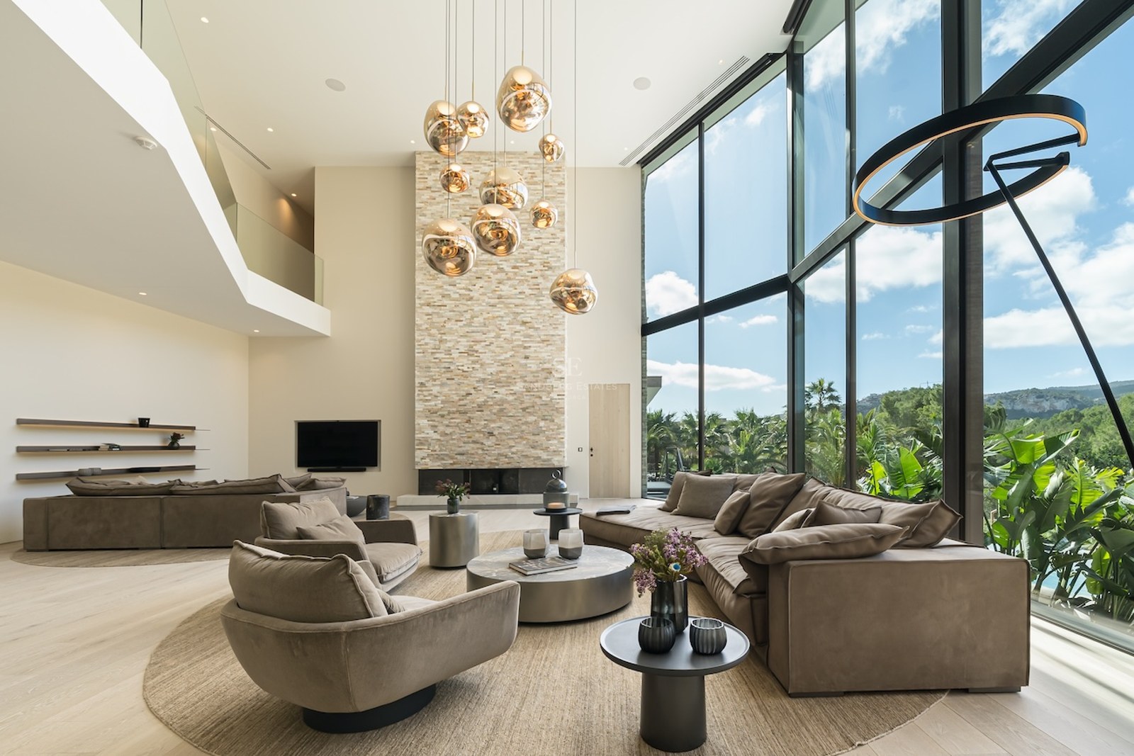 Un salon moderne à double hauteur avec cheminée en pierre, suspensions dorées et grandes baies vitrées sur le jardin.