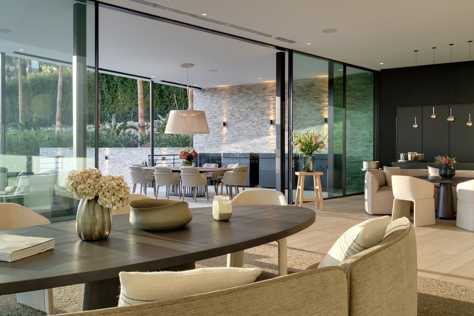 Elegante comedor con grandes ventanales de vidrio que abren a una terraza con pared de piedra y zona de estar exterior.