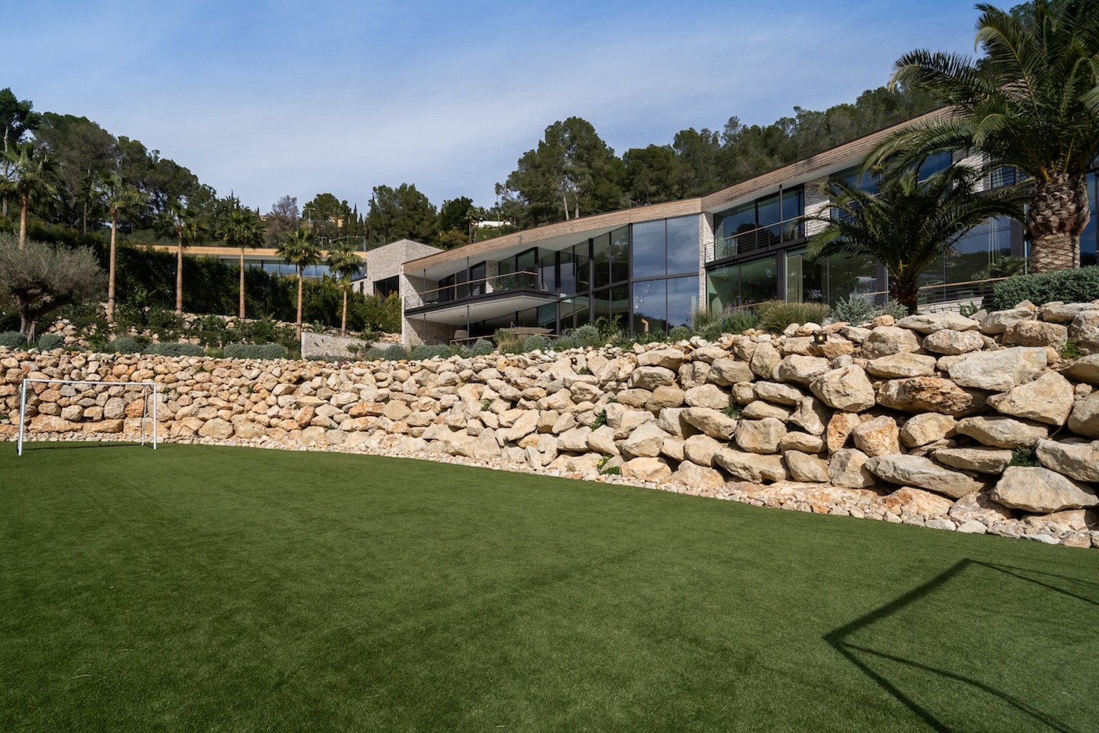 Façade de villa contemporaine avec baies vitrées derrière un mur de soutènement en pierre naturelle et pelouse verte.