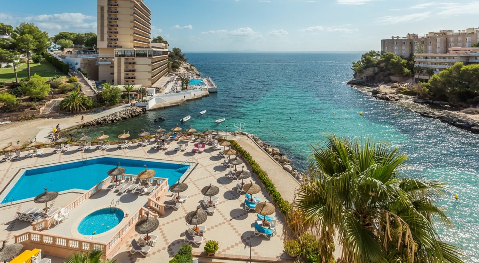 Vue aérienne d'une piscine avec chaises longues et parasols surplombant une baie turquoise et une côte rocheuse.