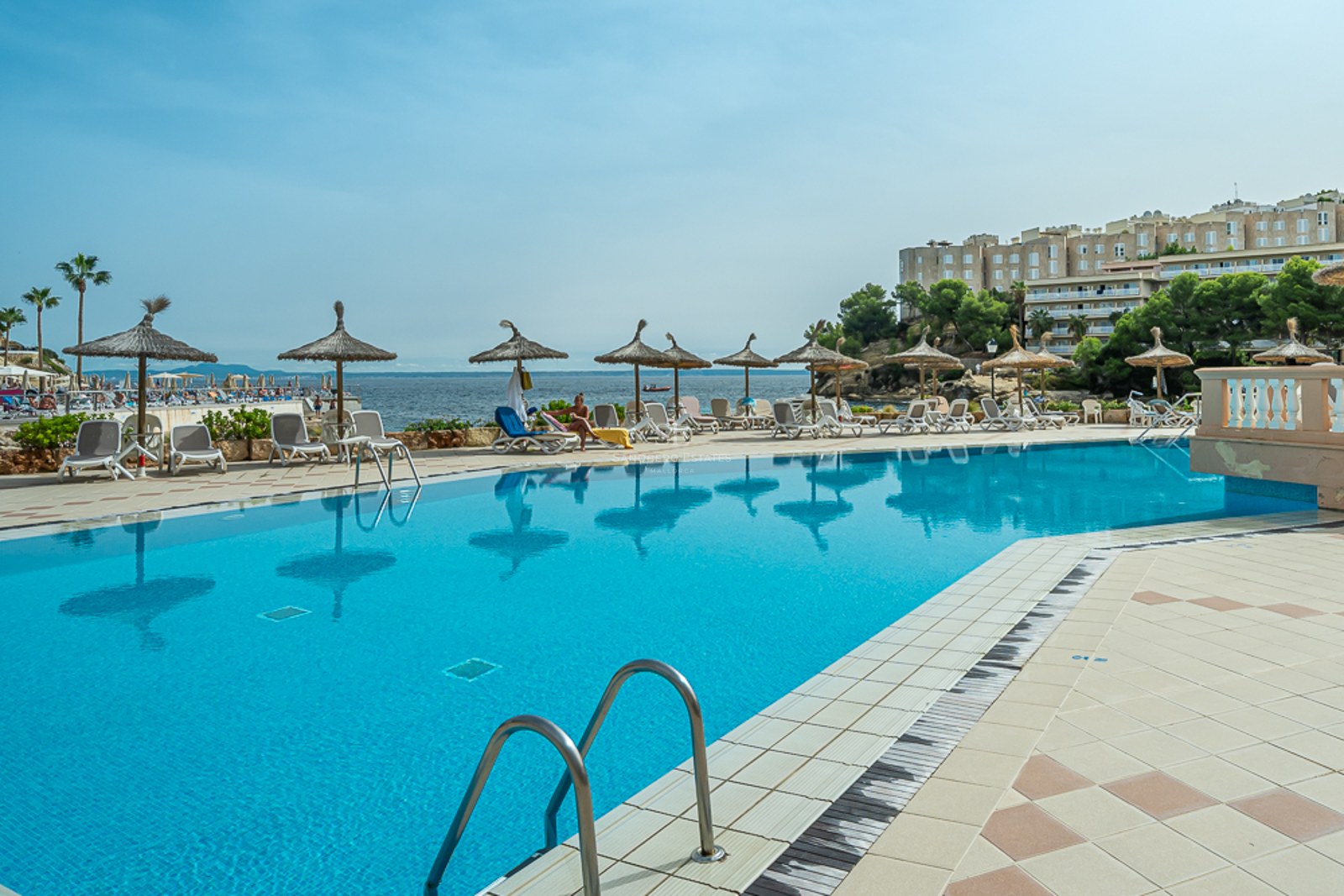 Grande piscine bleue entourée de transats et de parasols en paille avec vue sur la mer et les bâtiments côtiers.
