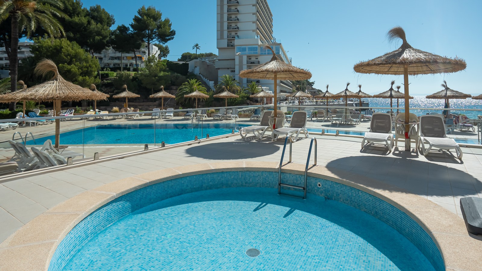 Espace piscine extérieur lumineux avec parasols en paille, transats blancs, balustrades en verre et vue sur la mer.