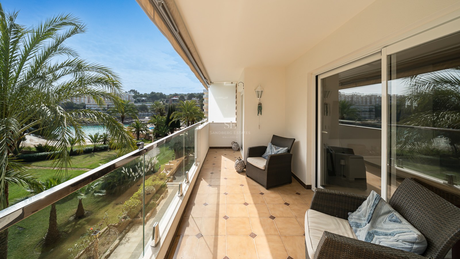 Balcon ensoleillé avec chaises en osier, garde-corps en verre et vue sur des palmiers et jardins.