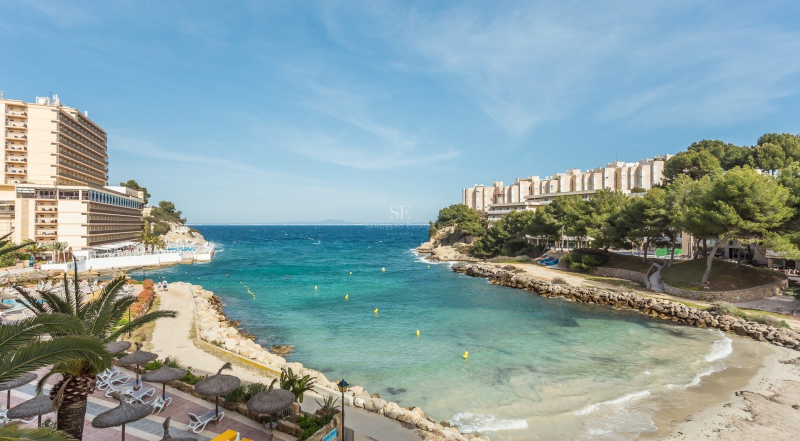 Vue en plongée d'une crique aux eaux turquoise avec une plage, des sentiers en pierre et des immeubles côtiers.
