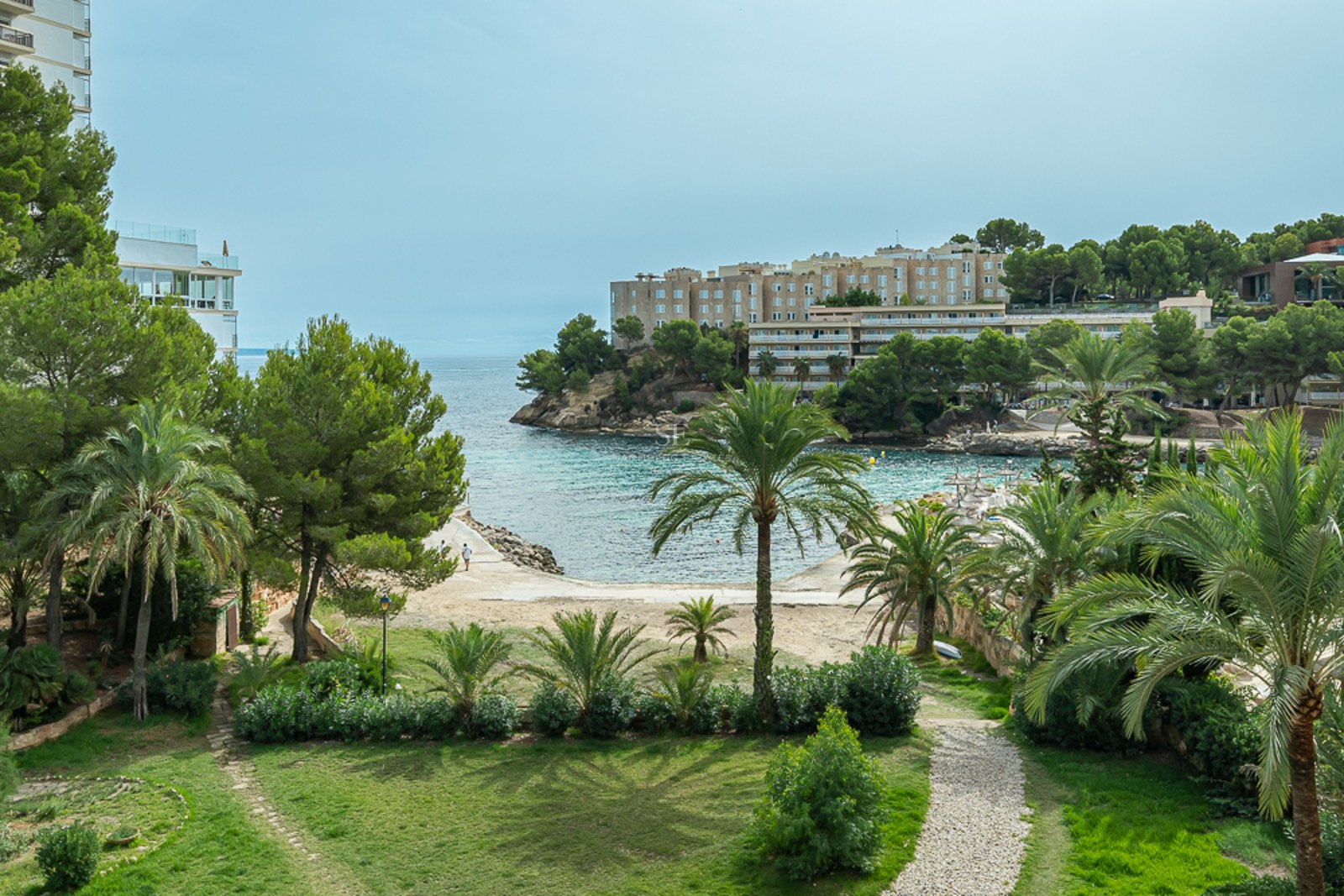 Un jardin luxuriant avec des palmiers et un chemin en pierre surplombant une crique méditerranéenne aux eaux turquoises.