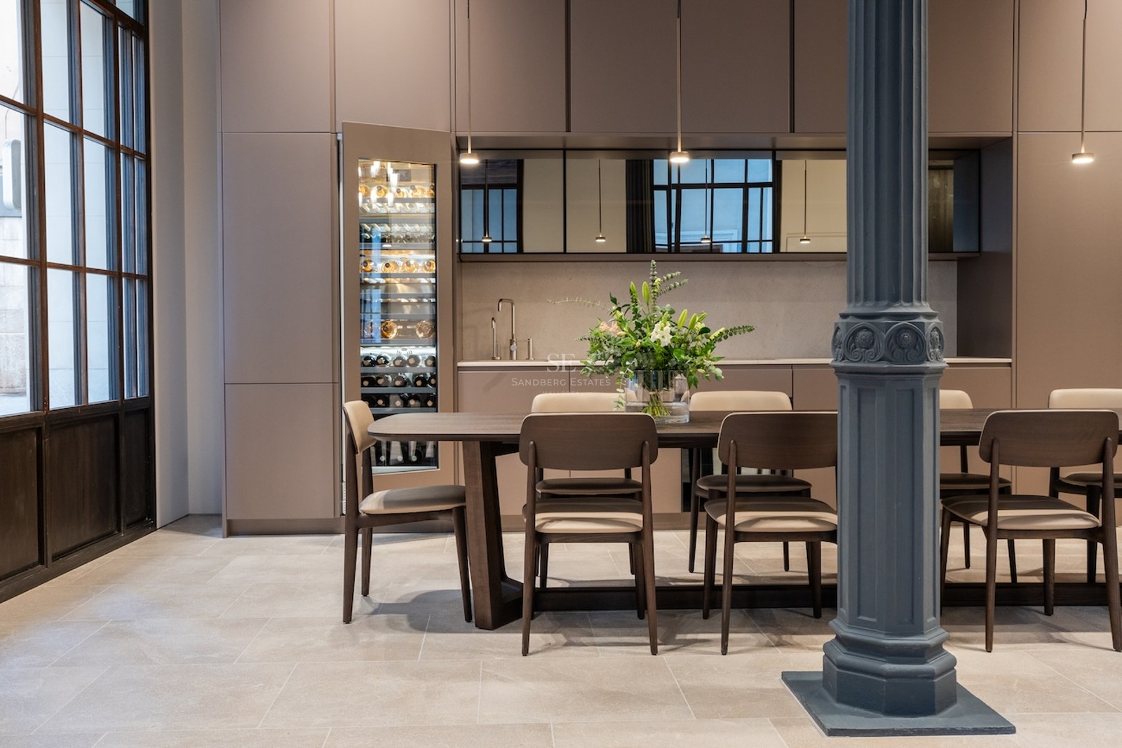 Comedor moderno con mesa de madera, sillas beige, columna industrial azul y vinoteca de cristal integrada.