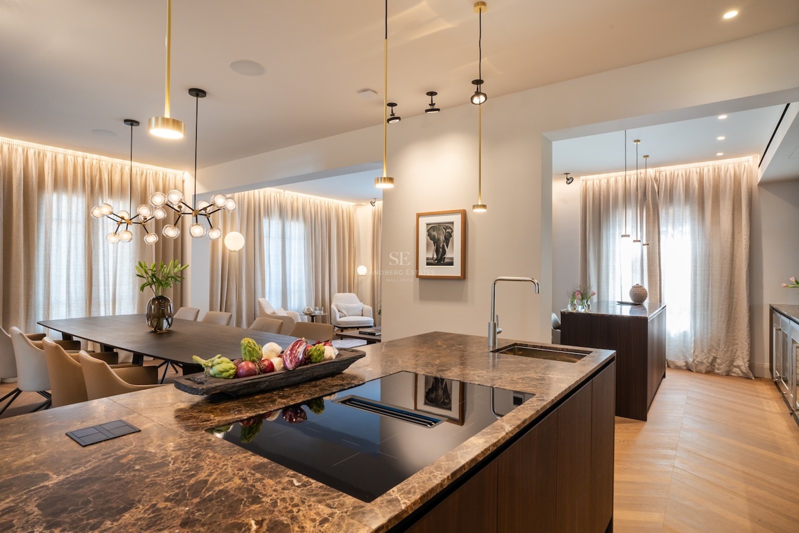 Vista desde una isla de cocina de mármol hacia una gran mesa de comedor y zona de estar con iluminación de diseño.