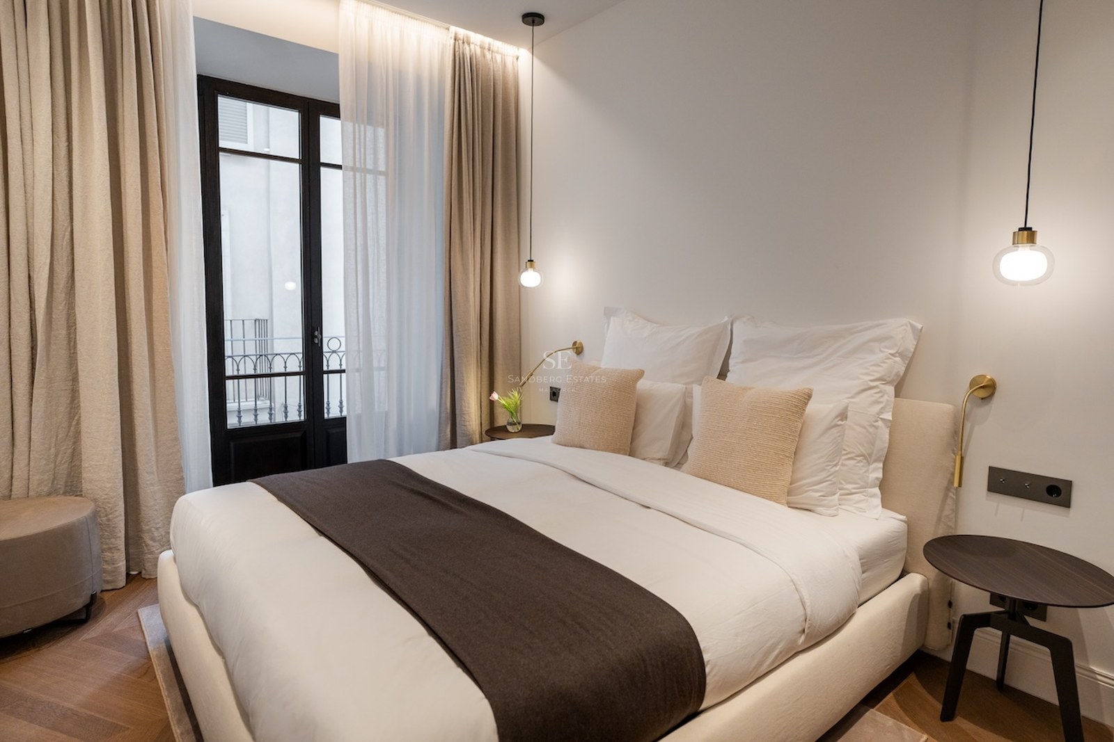 Dormitorio luminoso con cama doble, suelos de madera en espiga, lámparas colgantes y balcón.