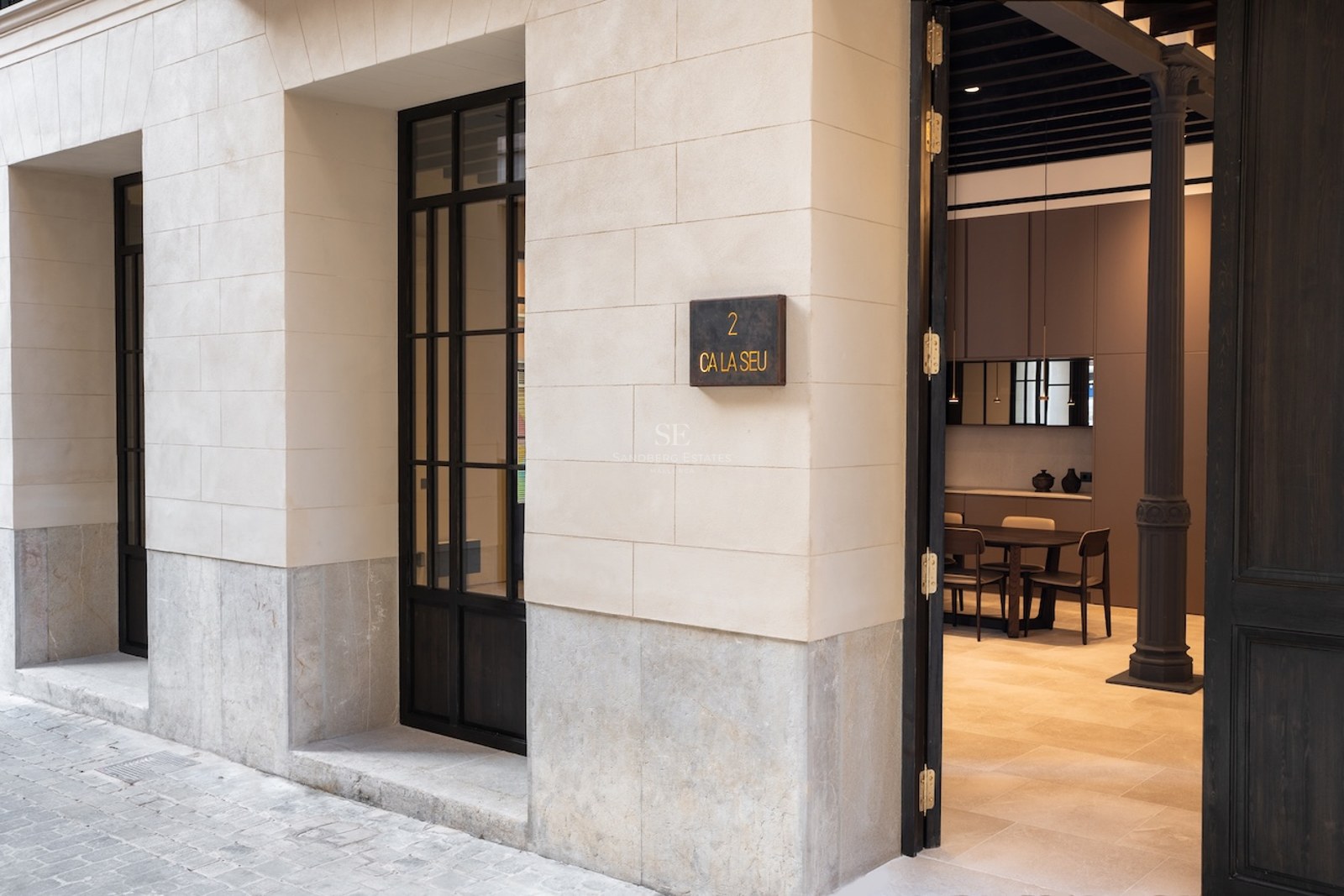 Primer plano de la entrada de un edificio moderno con bloques de piedra crema, puerta de metal negro y placa C'an Seu 2.