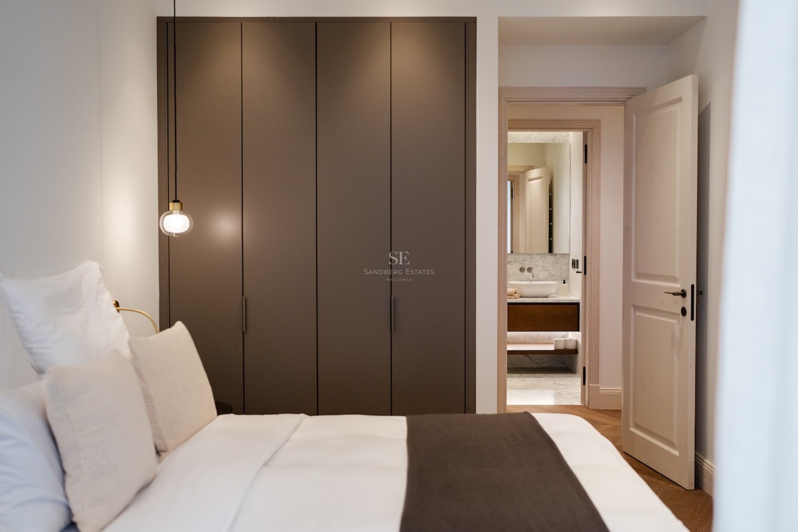 Dormitorio moderno con armario taupe, ropa de cama blanca y vista a un baño de mármol a través de una puerta abierta.