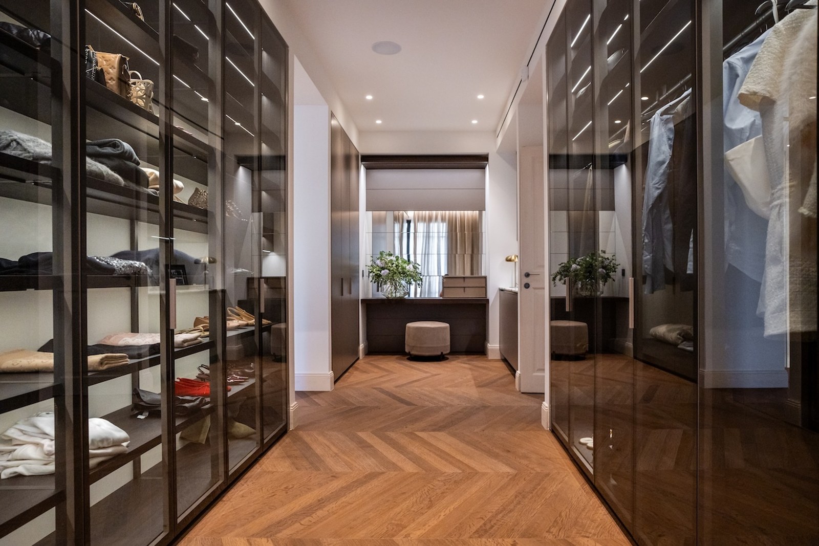 Elegante vestidor con armarios de cristal, suelos de madera en espiga y un tocador al fondo.