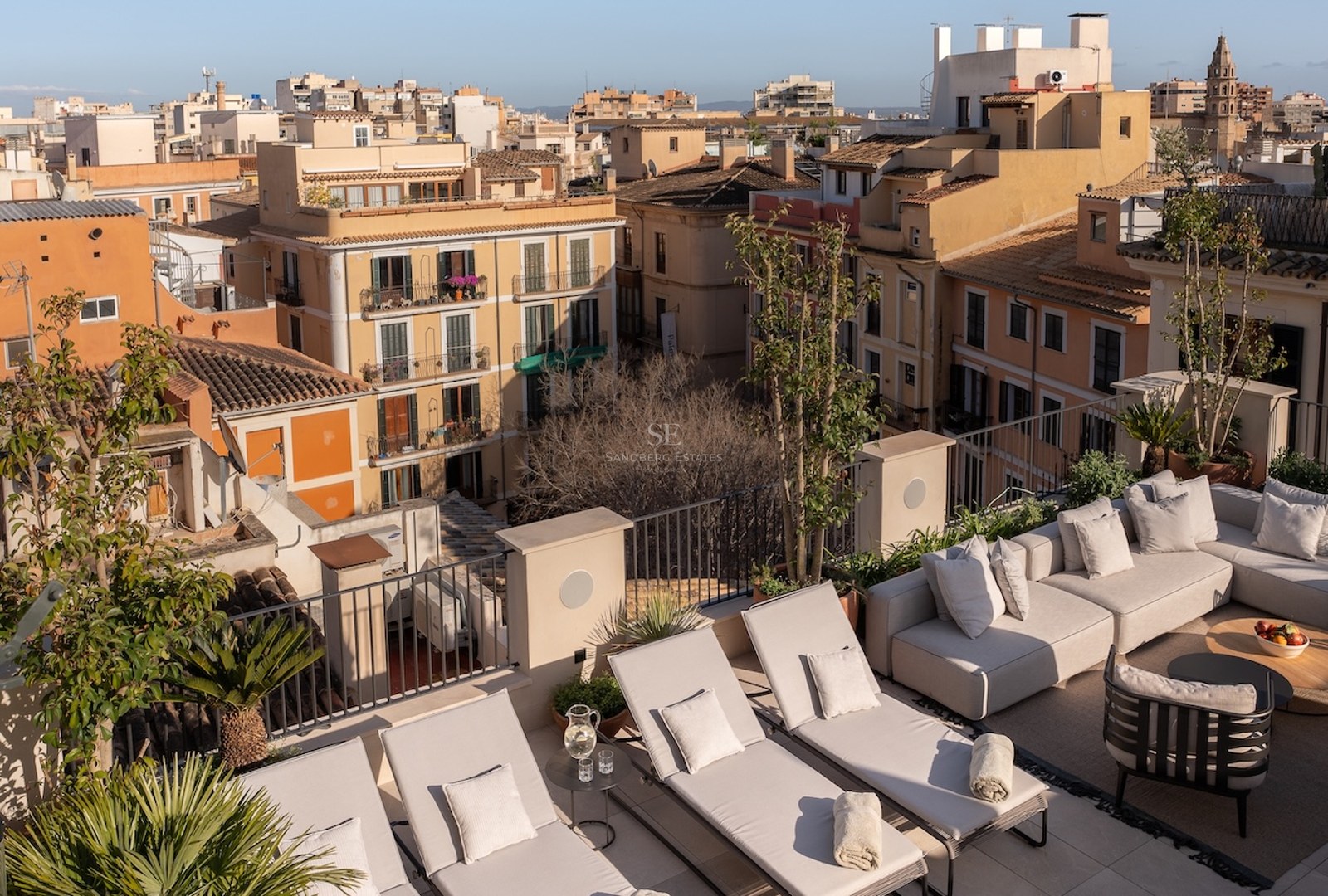 Terraza en la azotea con tumbonas beige, sofá esquinero y plantas con vistas a una ciudad histórica mediterránea.