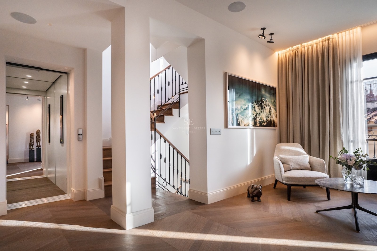 Pasillo de lujo con suelo de madera en espiga, ascensor privado, escalera y un sillón de diseño blanco.