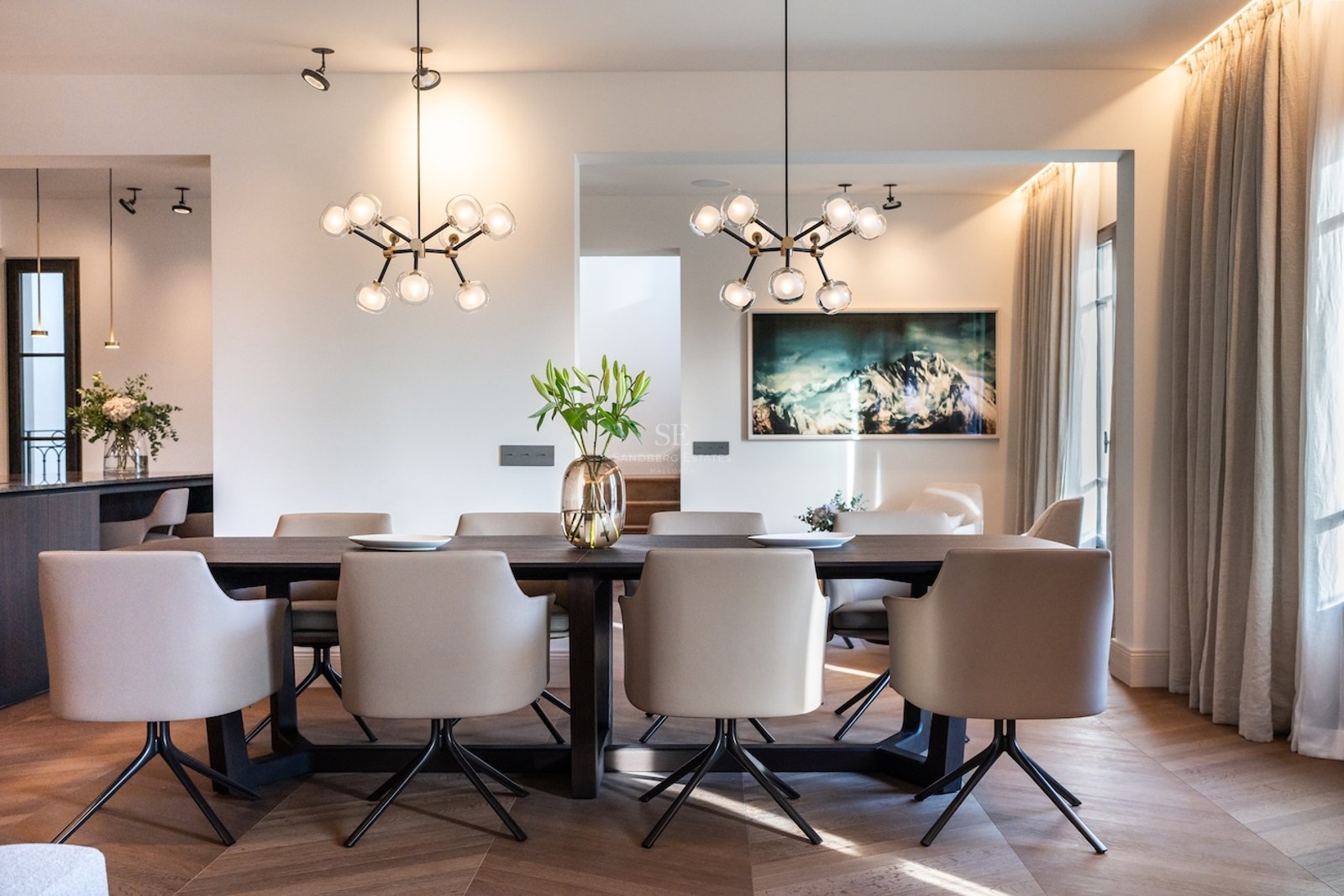 Comedor elegante con mesa de madera oscura, sillas beige, lámparas de esfera y cuadro de montaña.
