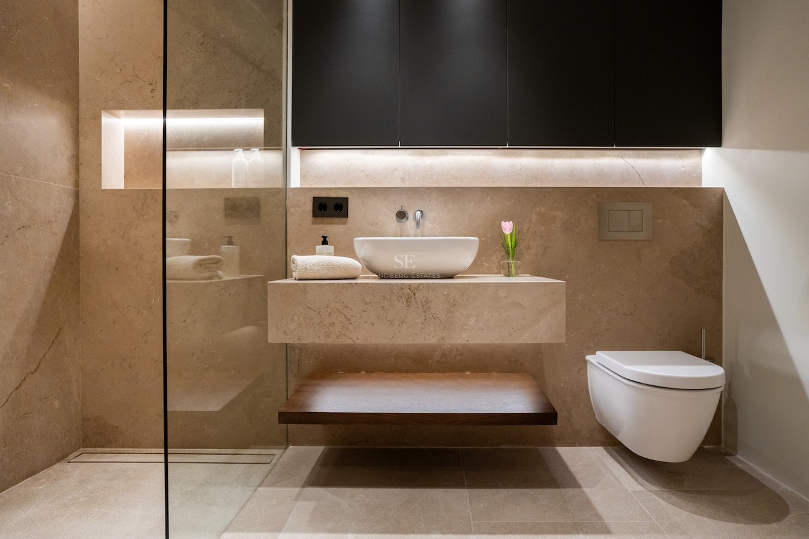 Baño moderno con paredes de piedra beige, lavabo flotante de mármol e iluminación LED integrada.