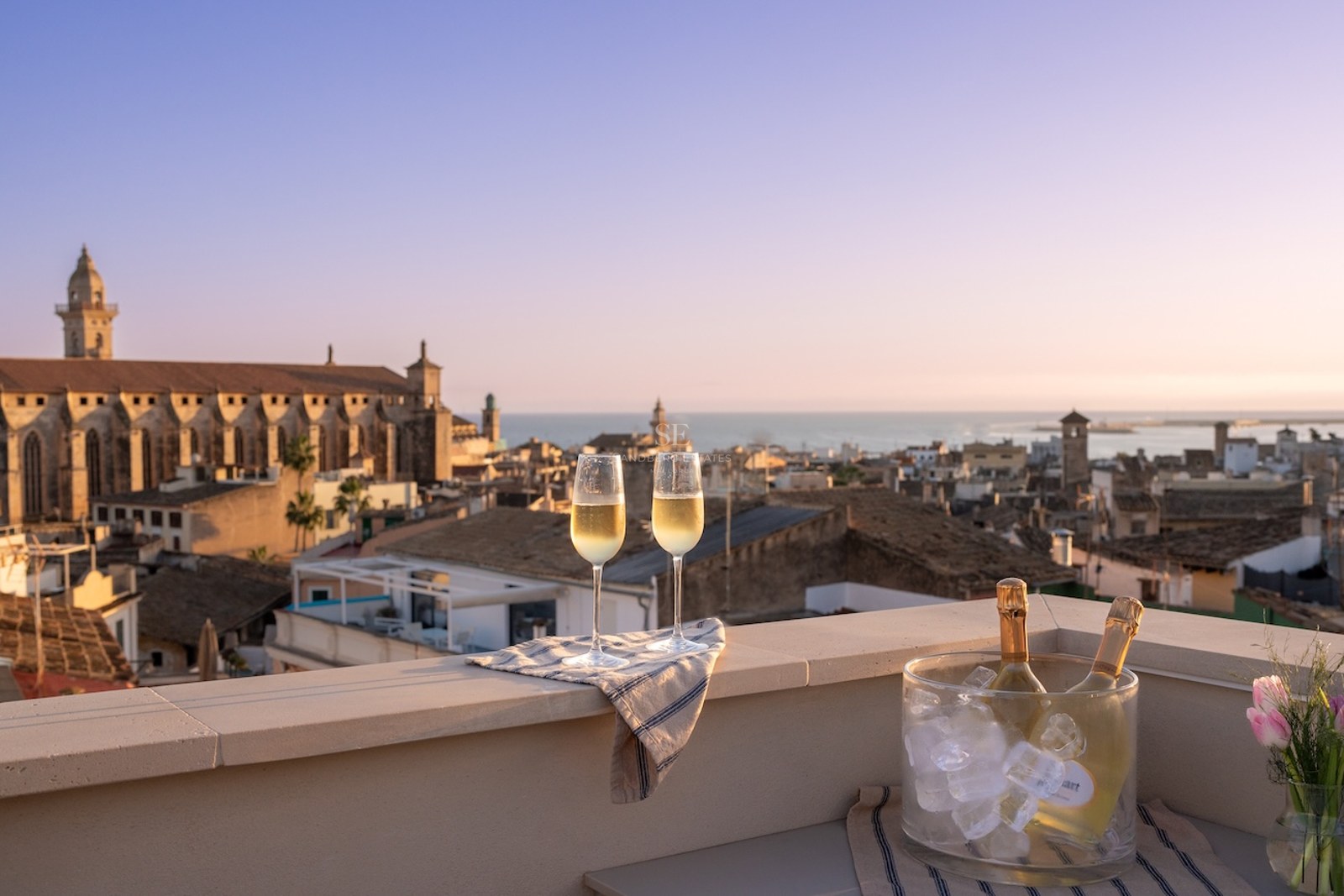 Dos copas de champán y una cubitera en una azotea con vistas a una ciudad costera histórica al atardecer.