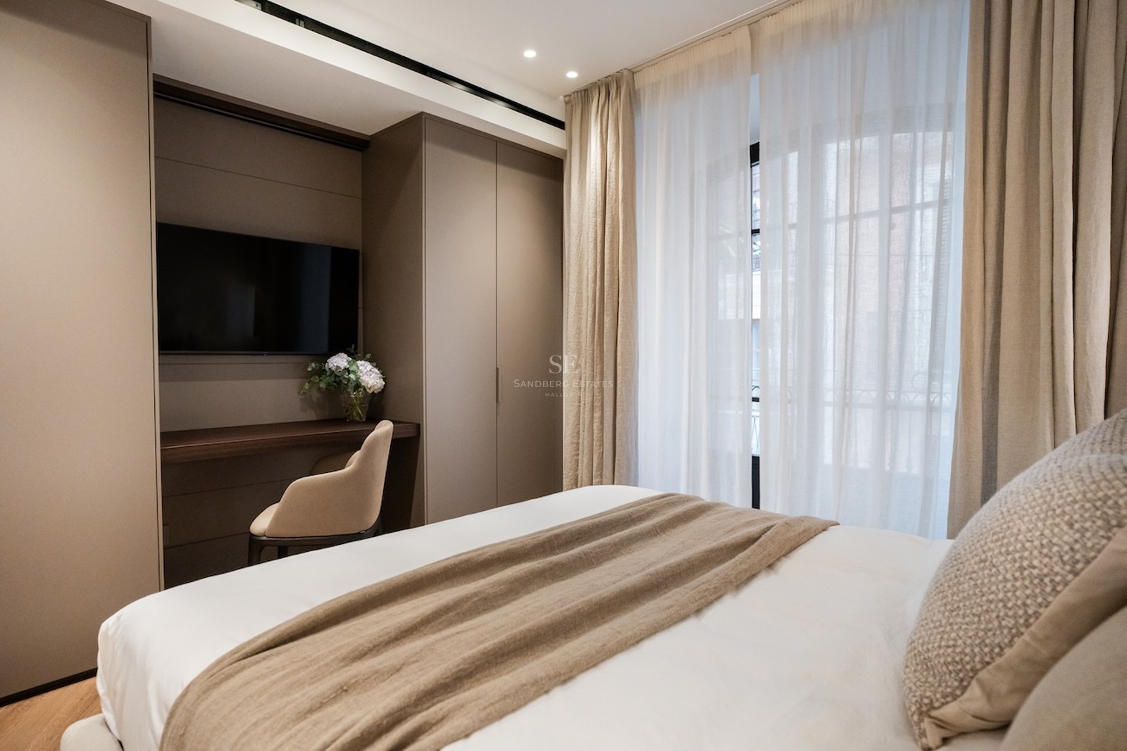 Dormitorio moderno con cama blanca, manta beige, escritorio integrado con TV y grandes ventanales con cortinas.