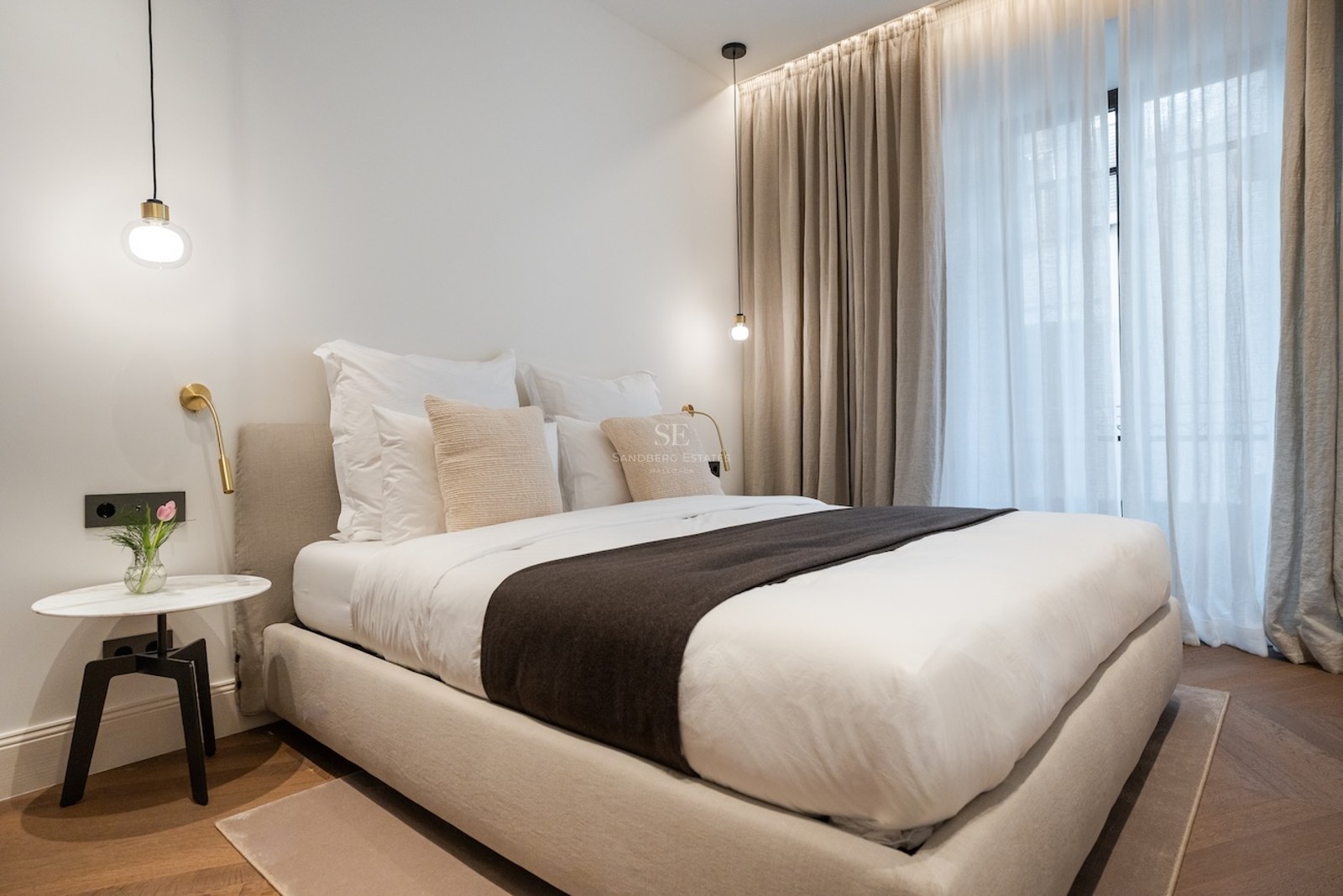 Dormitorio principal moderno con cama beige, mesita de mármol, suelo de parqué y cortinas de techo a suelo.