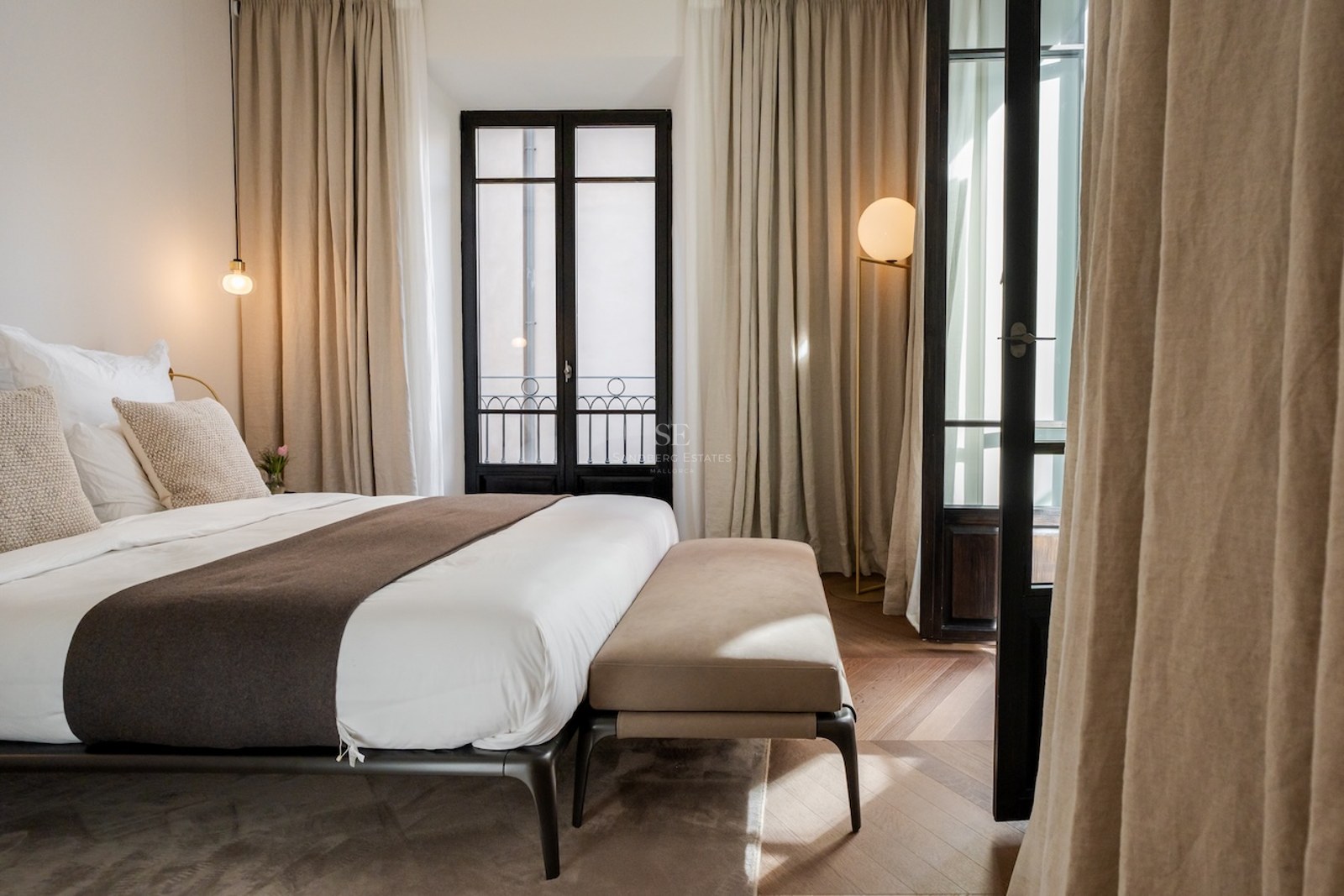 Dormitorio moderno con cama grande, cortinas de lino beige, suelo de madera en espiga y puertas de cristal negras.