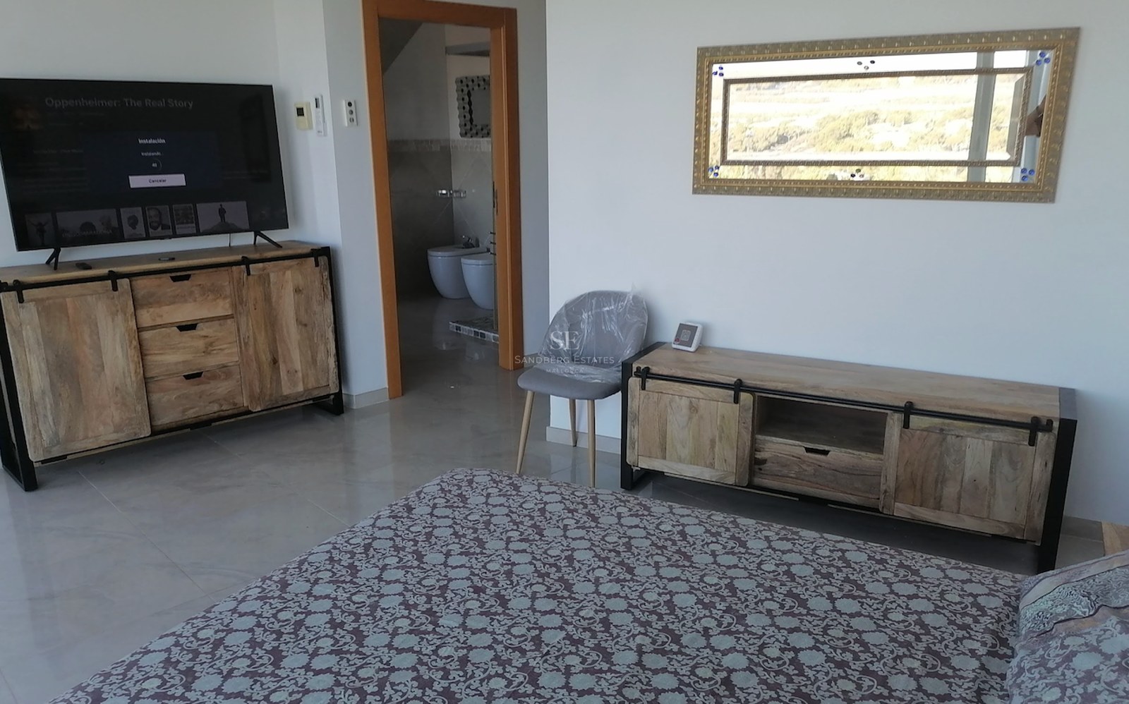 Modern bedroom featuring rustic wood media consoles, a large TV, and a view into a private en-suite bathroom.