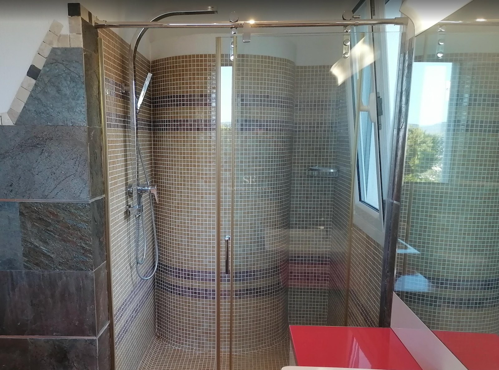 A walk-in glass shower enclosure featuring beige mosaic tiles, chrome fixtures, and a curved interior wall.