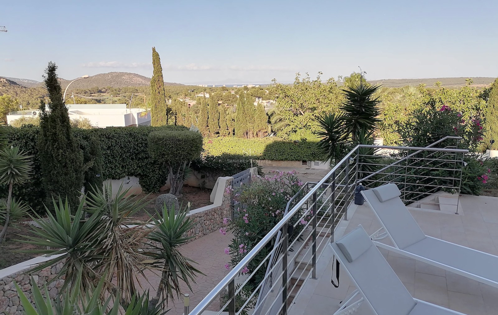 A sun-drenched terrace with modern loungers and stainless steel railings overlooking a lush Mediterranean garden and distant sea.
