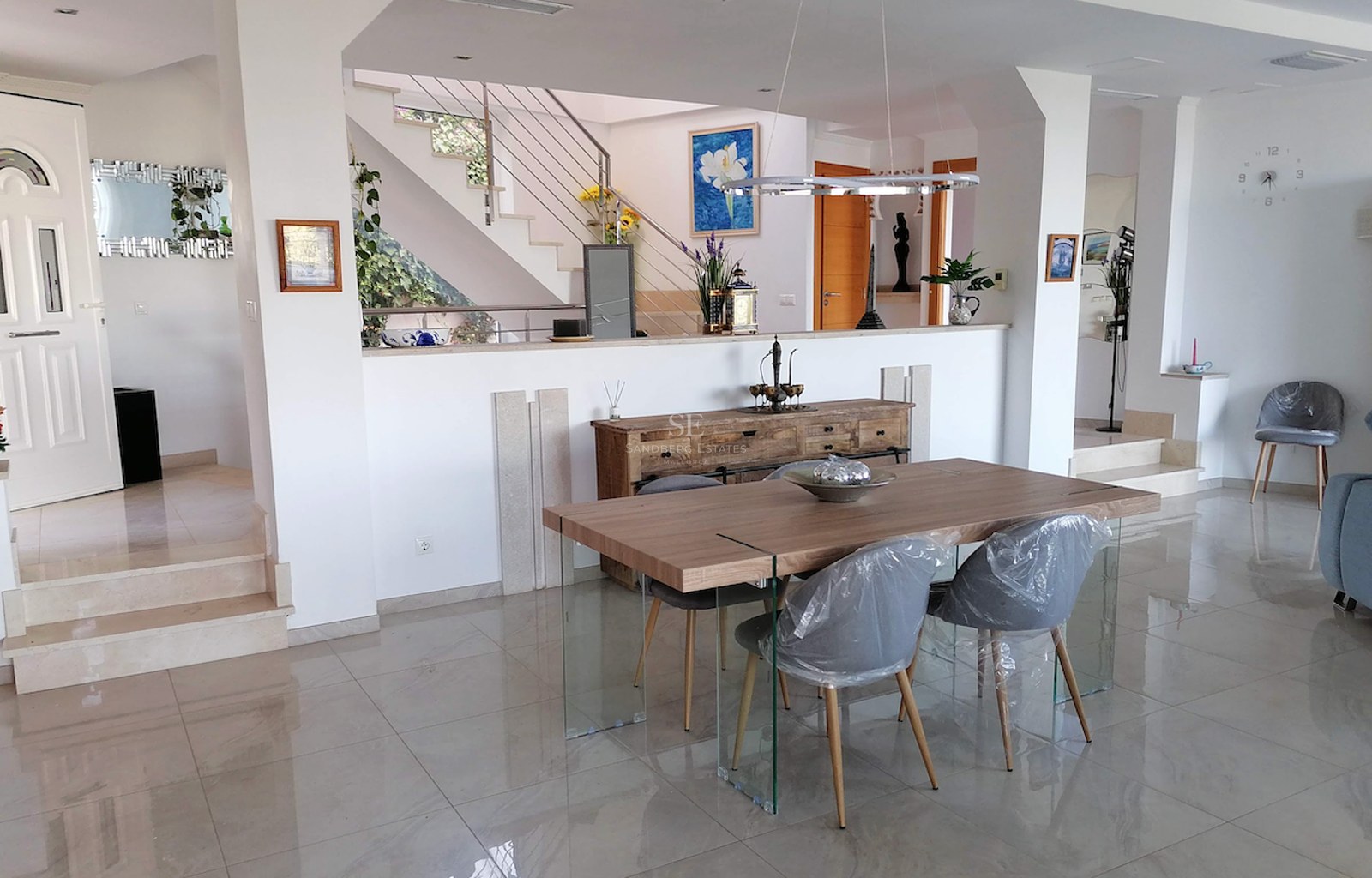 Modern dining room with wooden table, glass legs, marble floors, and stairs in the background.