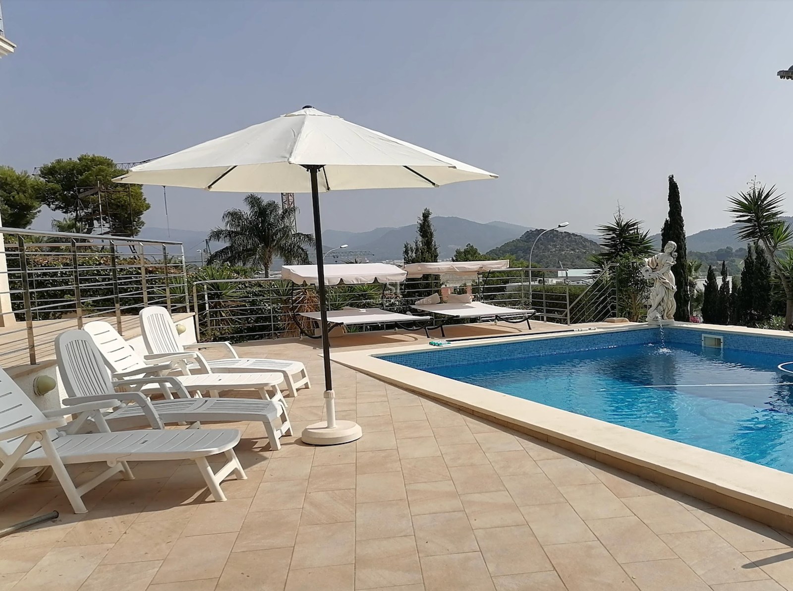 Sun-drenched pool area featuring white sun loungers, a large parasol, and a classic statue overlooking mountains.