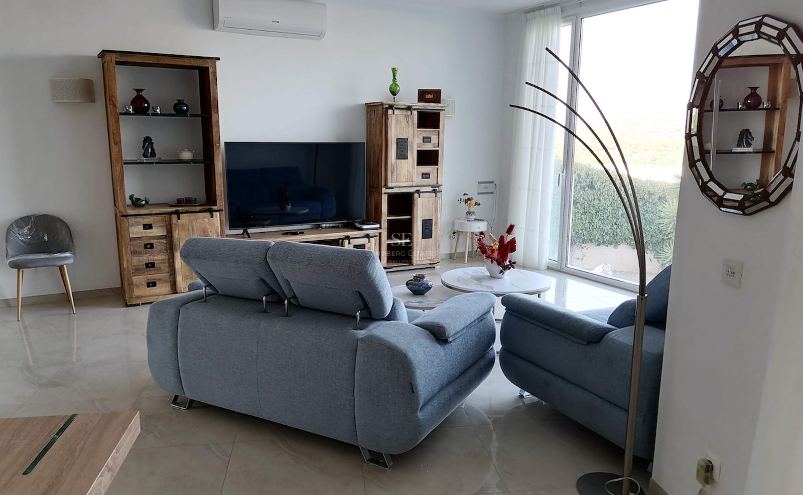 Modern living room featuring blue sofas, rustic wood cabinetry, polished tile flooring, and floor-to-ceiling windows.